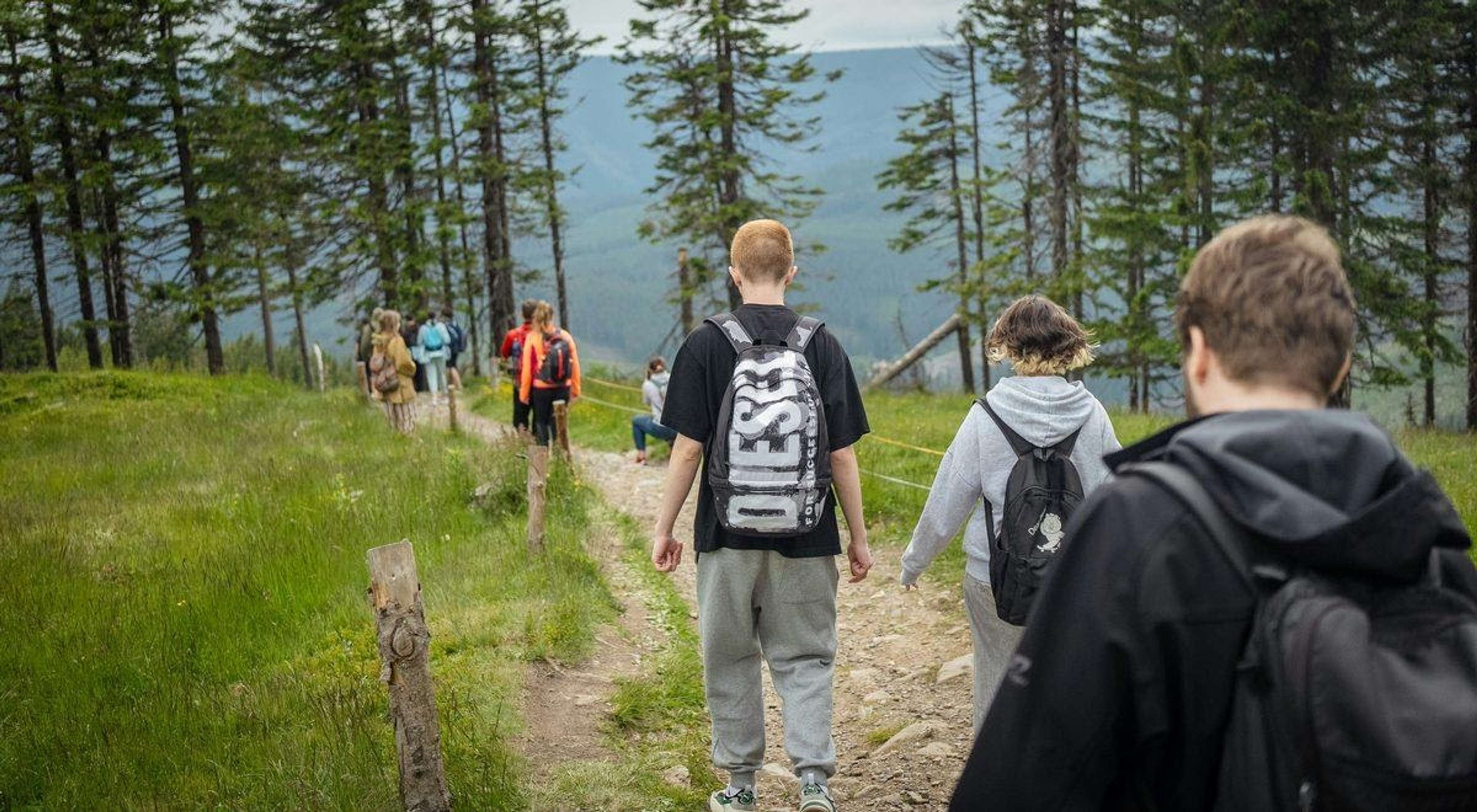 Ungdomar med ryggsäckar vandrar på en stig i skogen med berg i bakgrunden