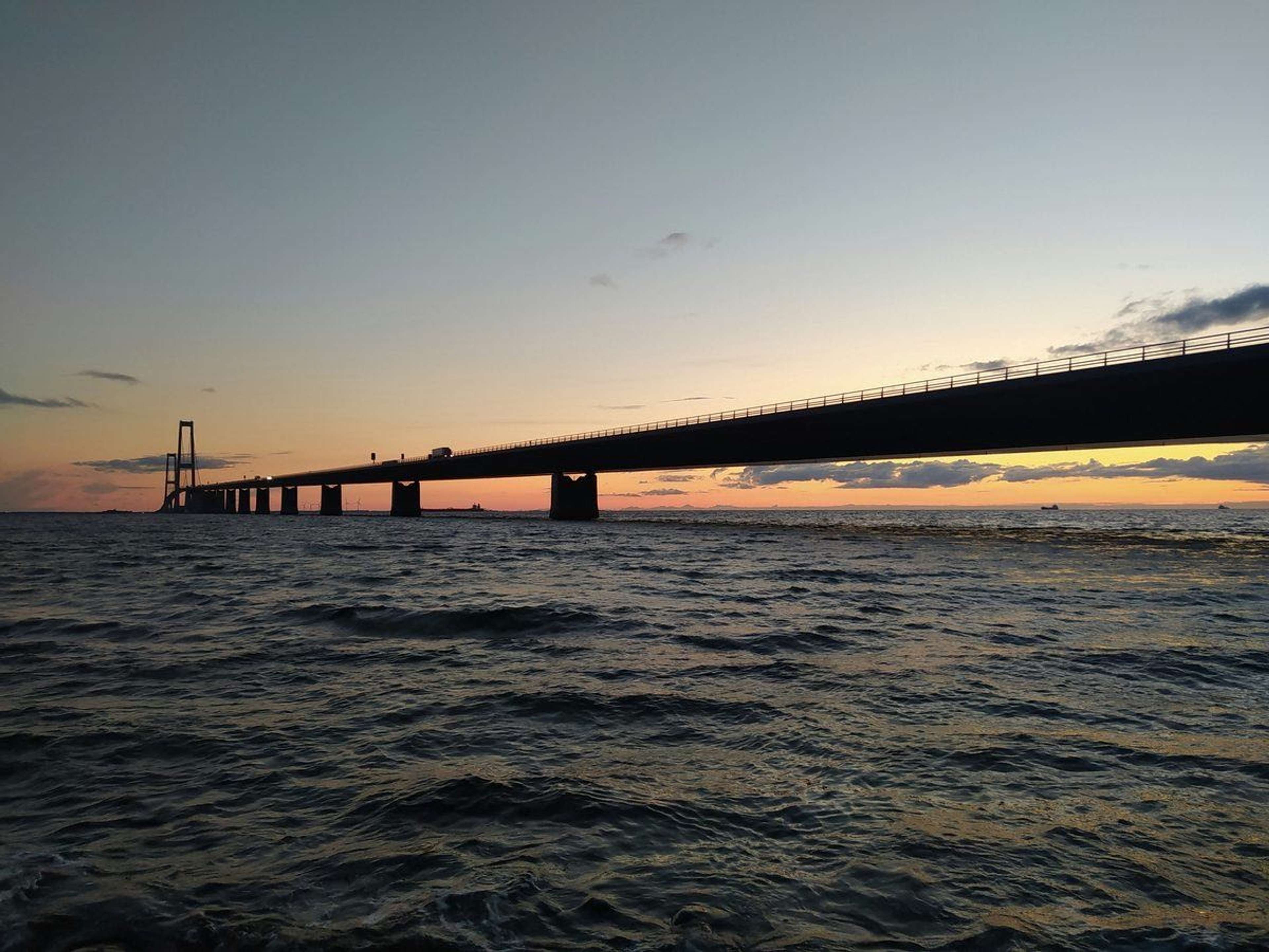 Lang bro over havet i solnedgang, set fra vandet med bølger i forgrunden