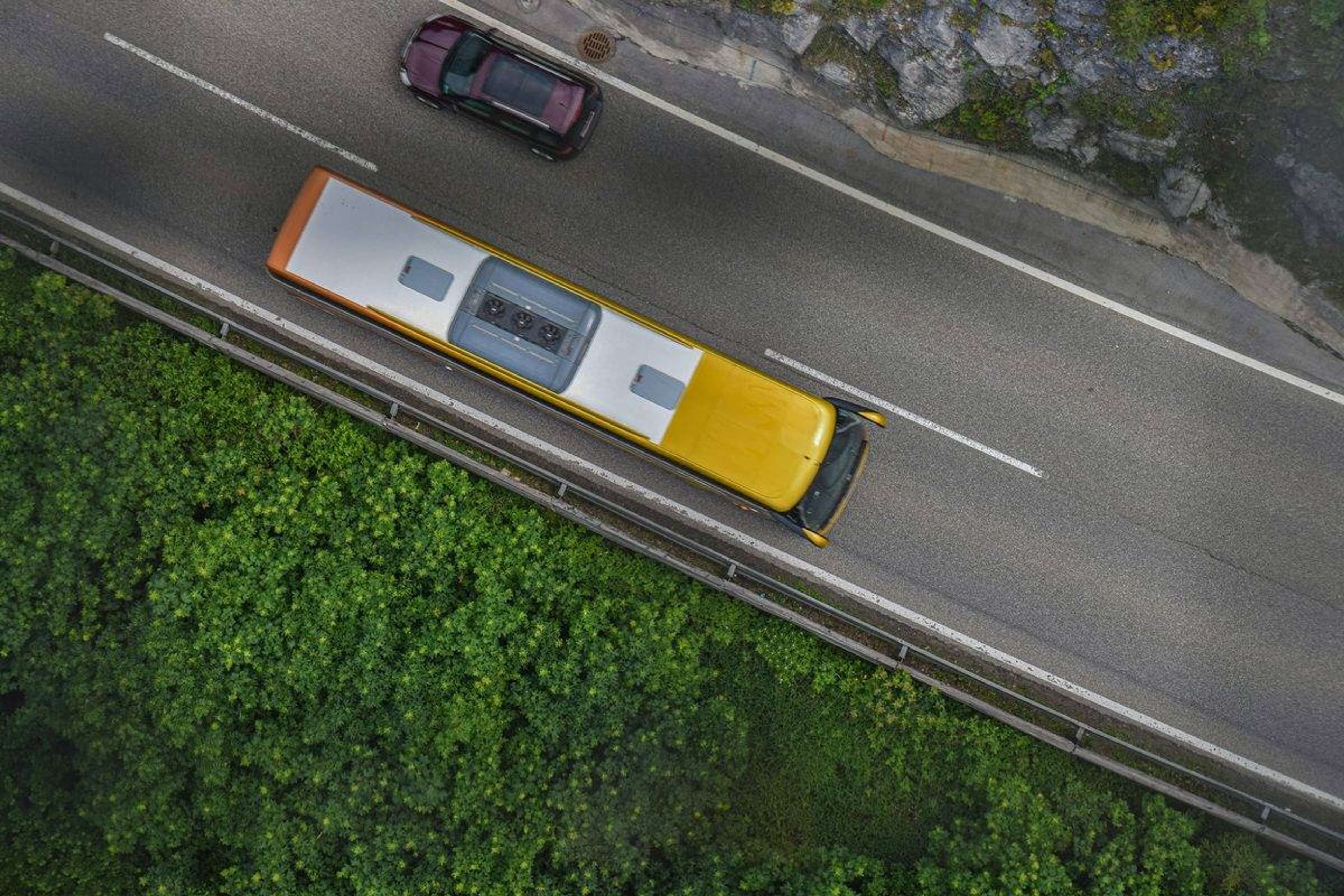 Gul buss med hvitt tak og en personbil sett ovenfra på en vei mellom fjellvegg og grønne trær