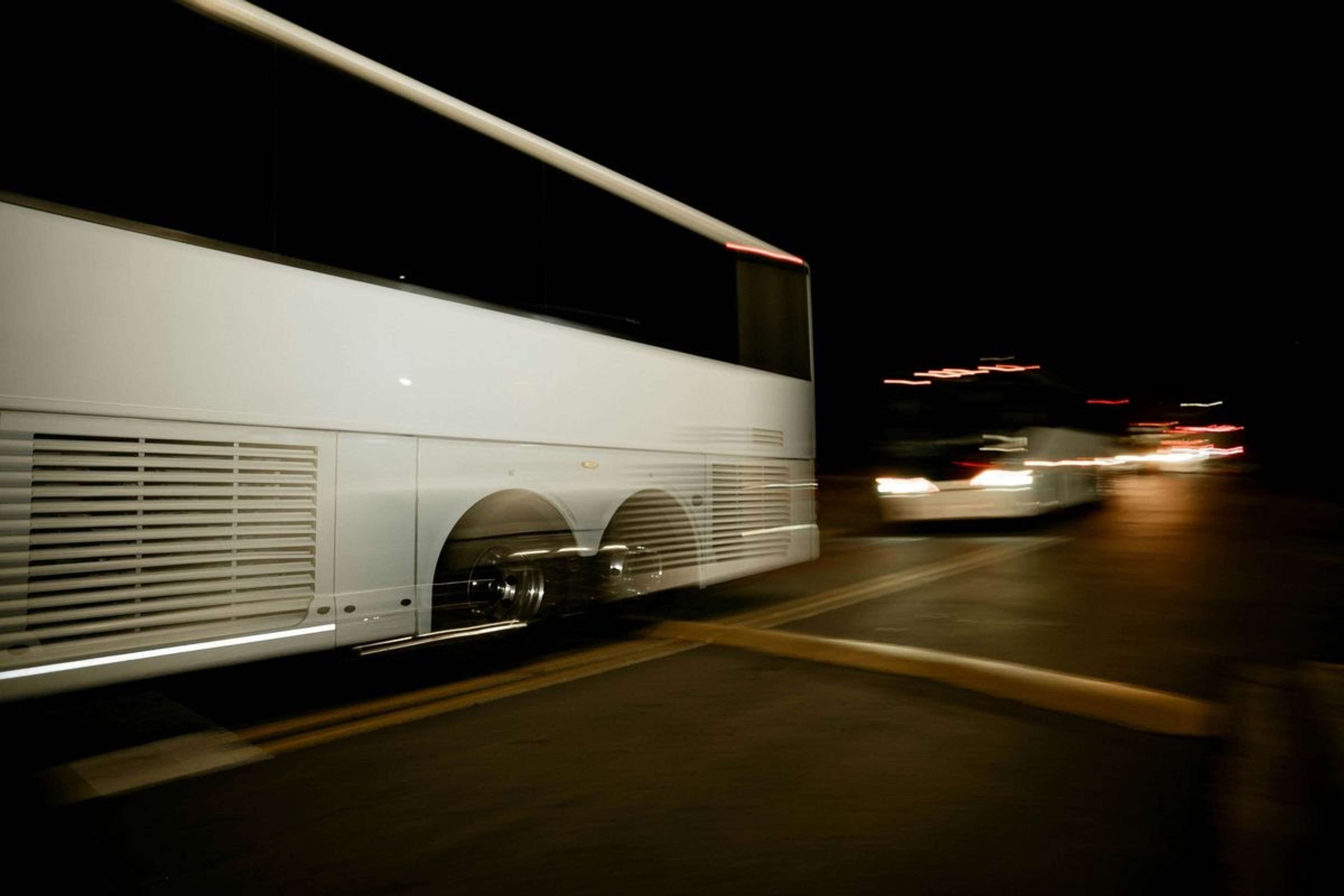 Vit turistbuss i rörelse på en mörk väg nattetid, med suddiga strålkastare från mötande trafik i bakgrunden