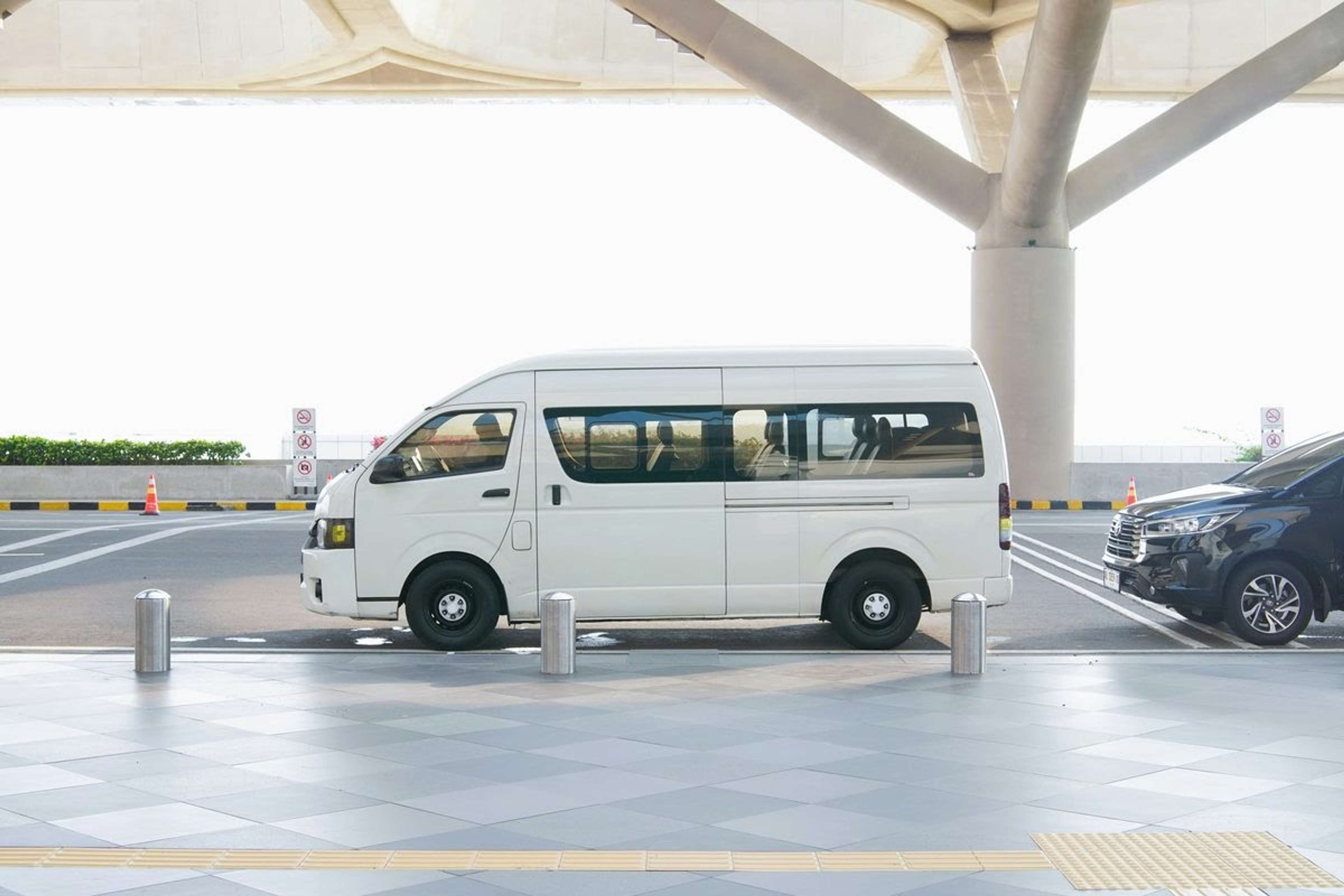 Hvit minibuss parkert foran en flyplassterminal, med en bil bak