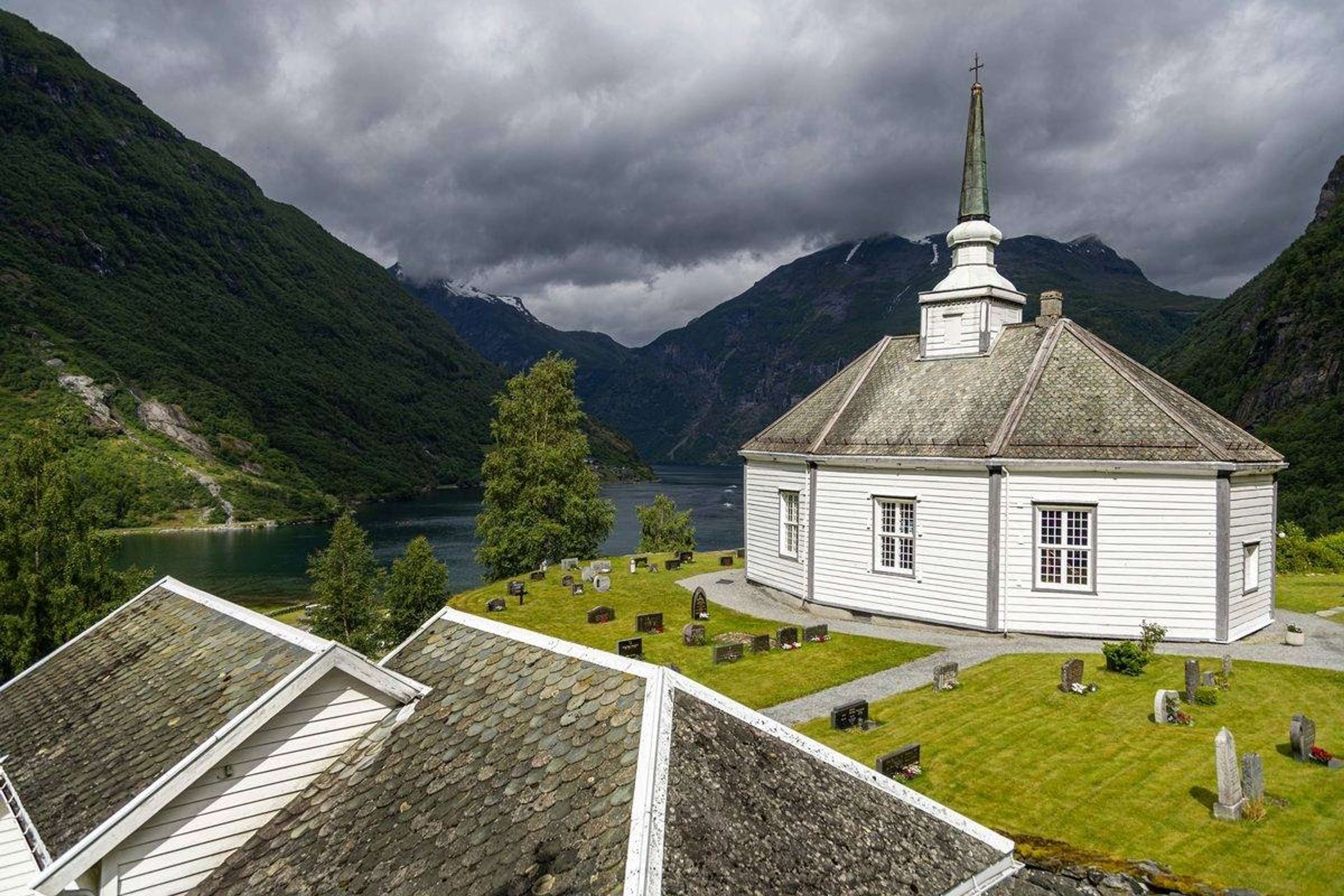 Hvit trekirke med kirkegård ved en fjord, med bratte fjell og mørke skyer bak