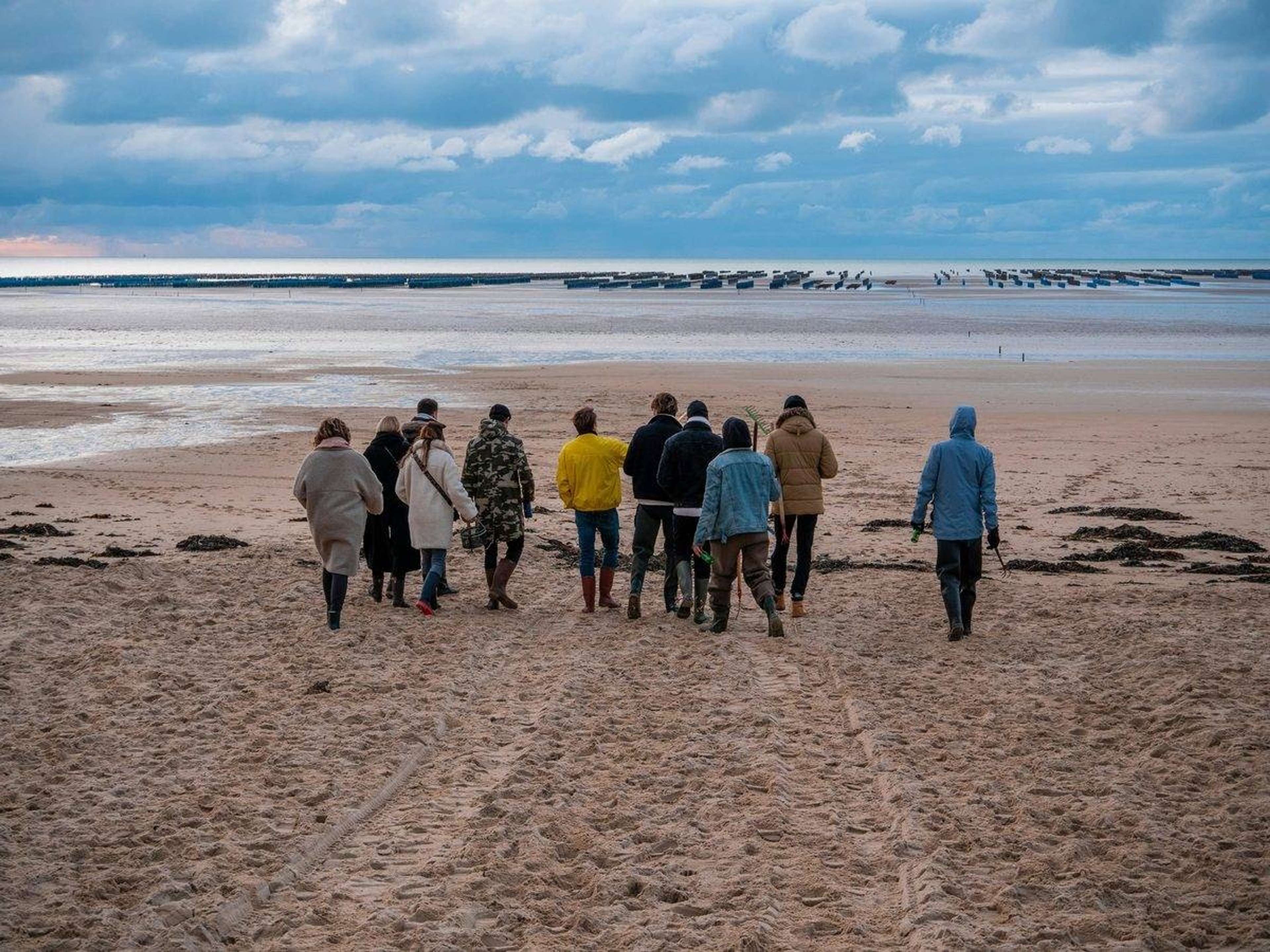 Gruppe mennesker går sammen på en bred strand ved lavvande under en overskyet himmel