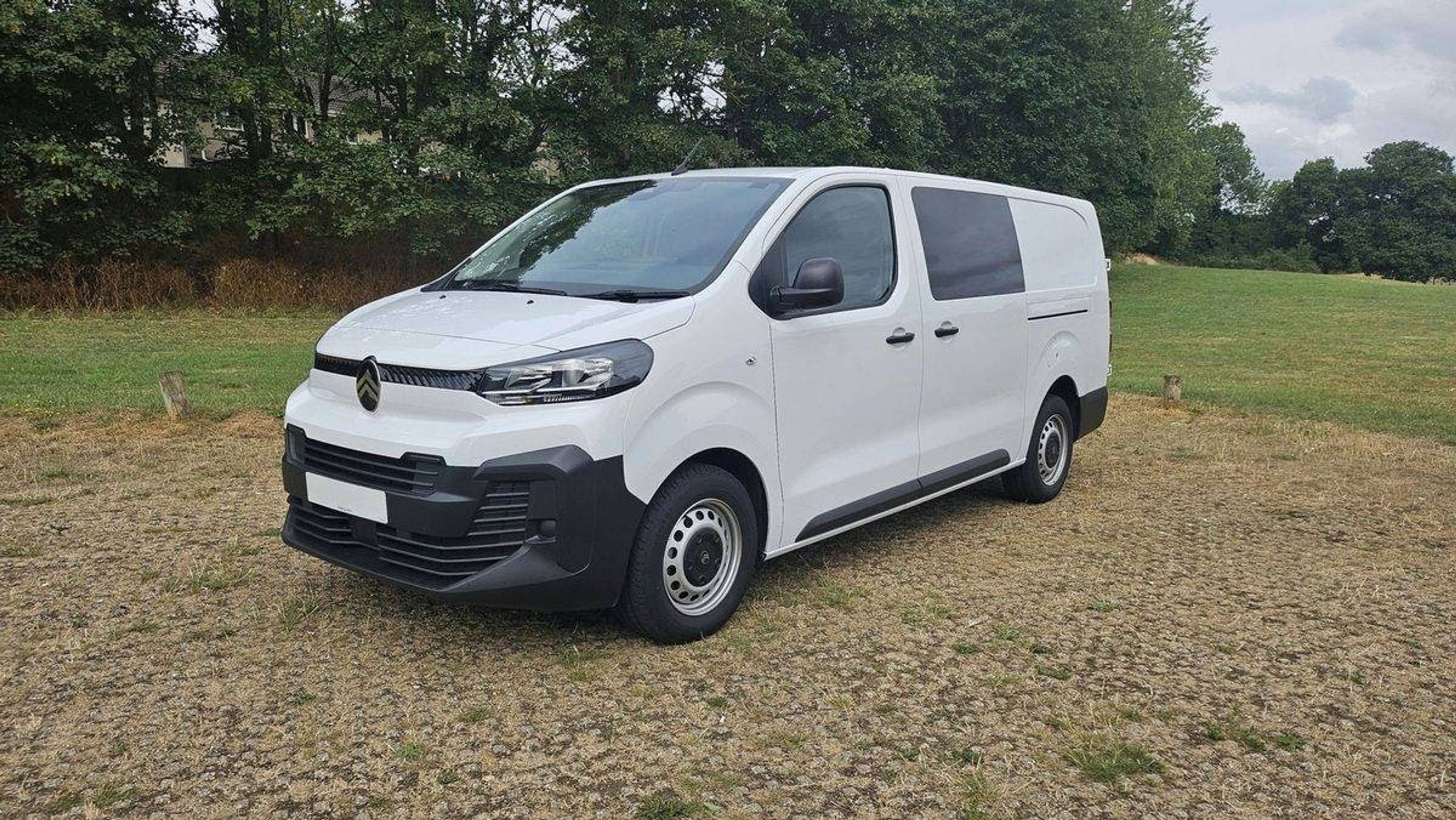 Valkoinen pieni pakettiauto pysäköitynä sorakentälle, taustalla nurmikkoa ja puita