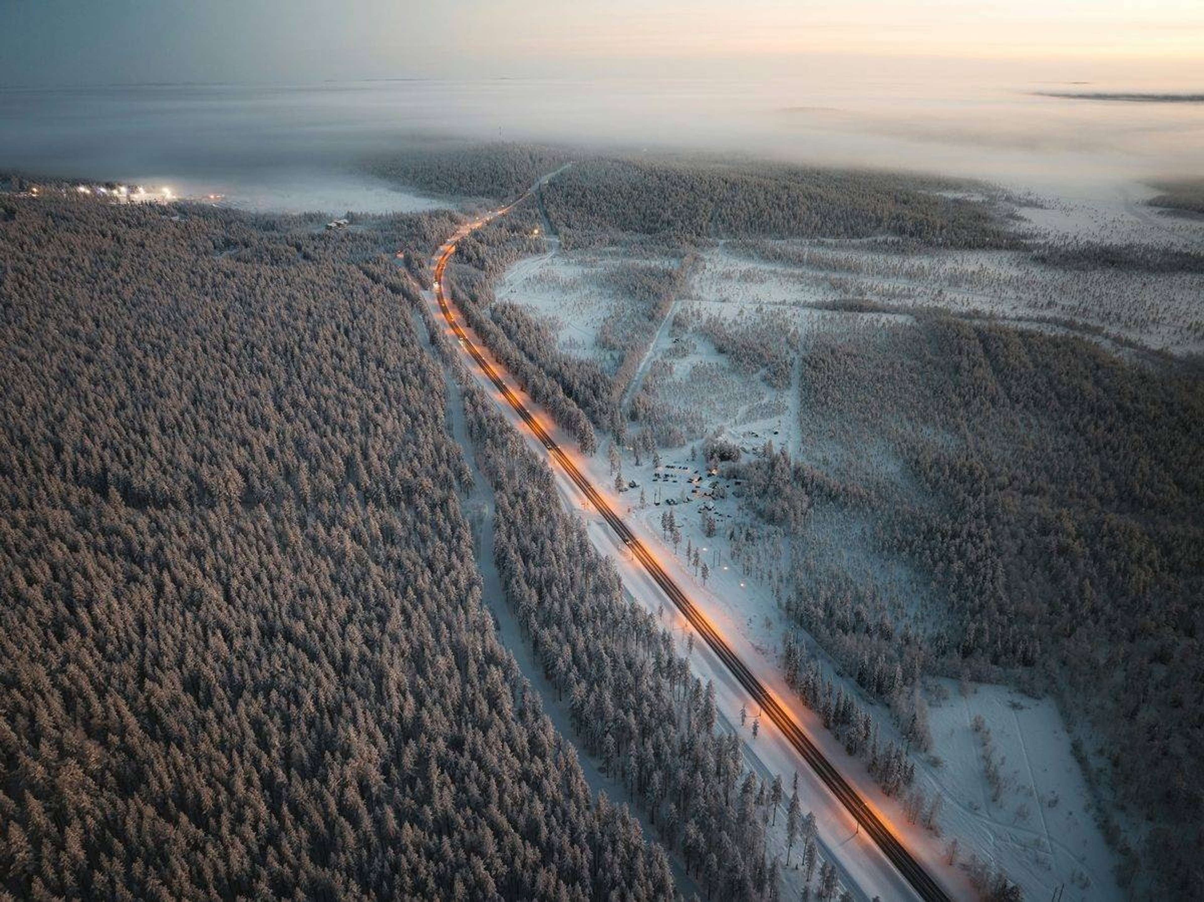 Ilmakuva valaistusta tiestä lumisen metsän halki talvisessa hämärässä