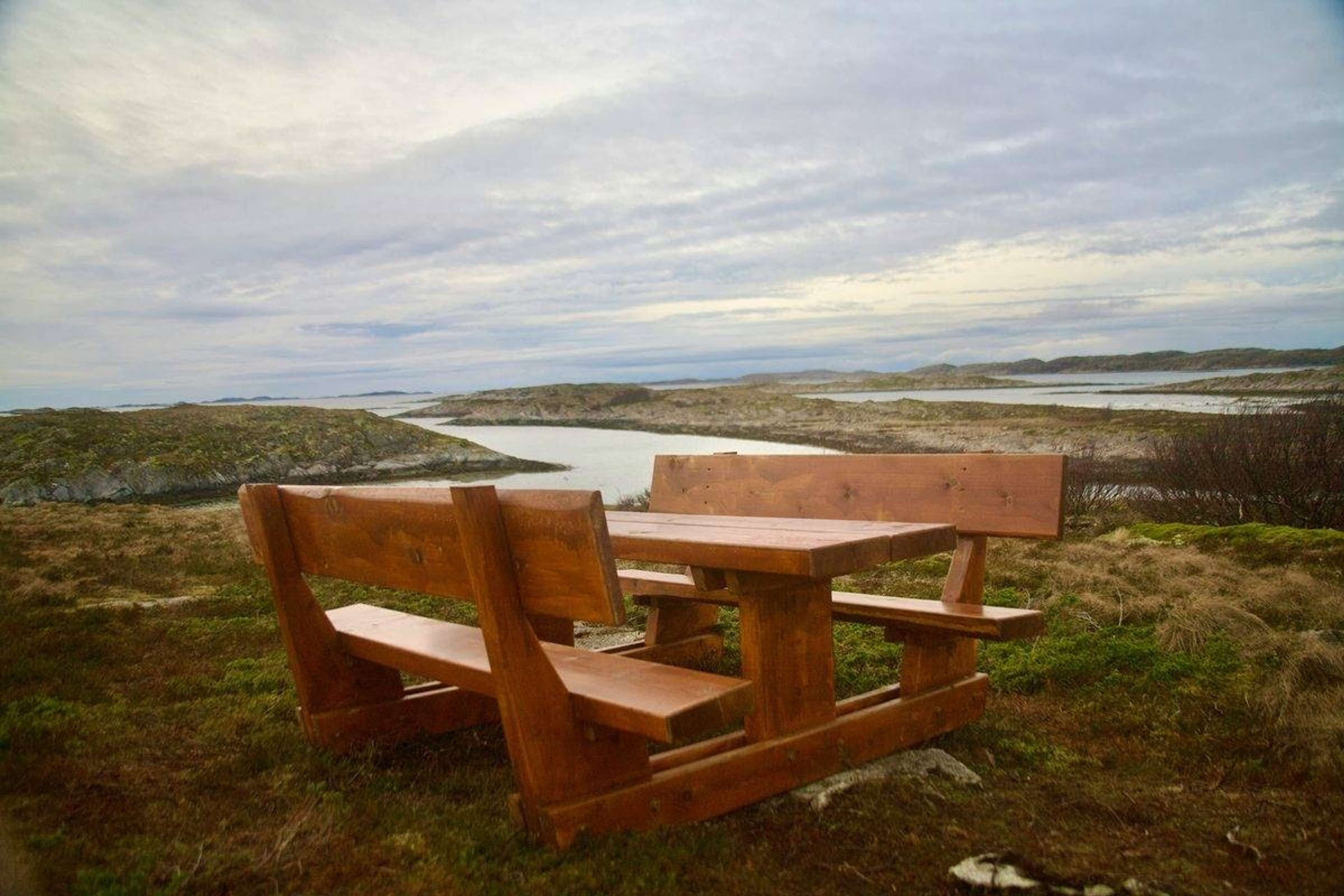 Piknikbord i tre på en gresskledd høyde med utsikt over vann og holmer under skyet himmel