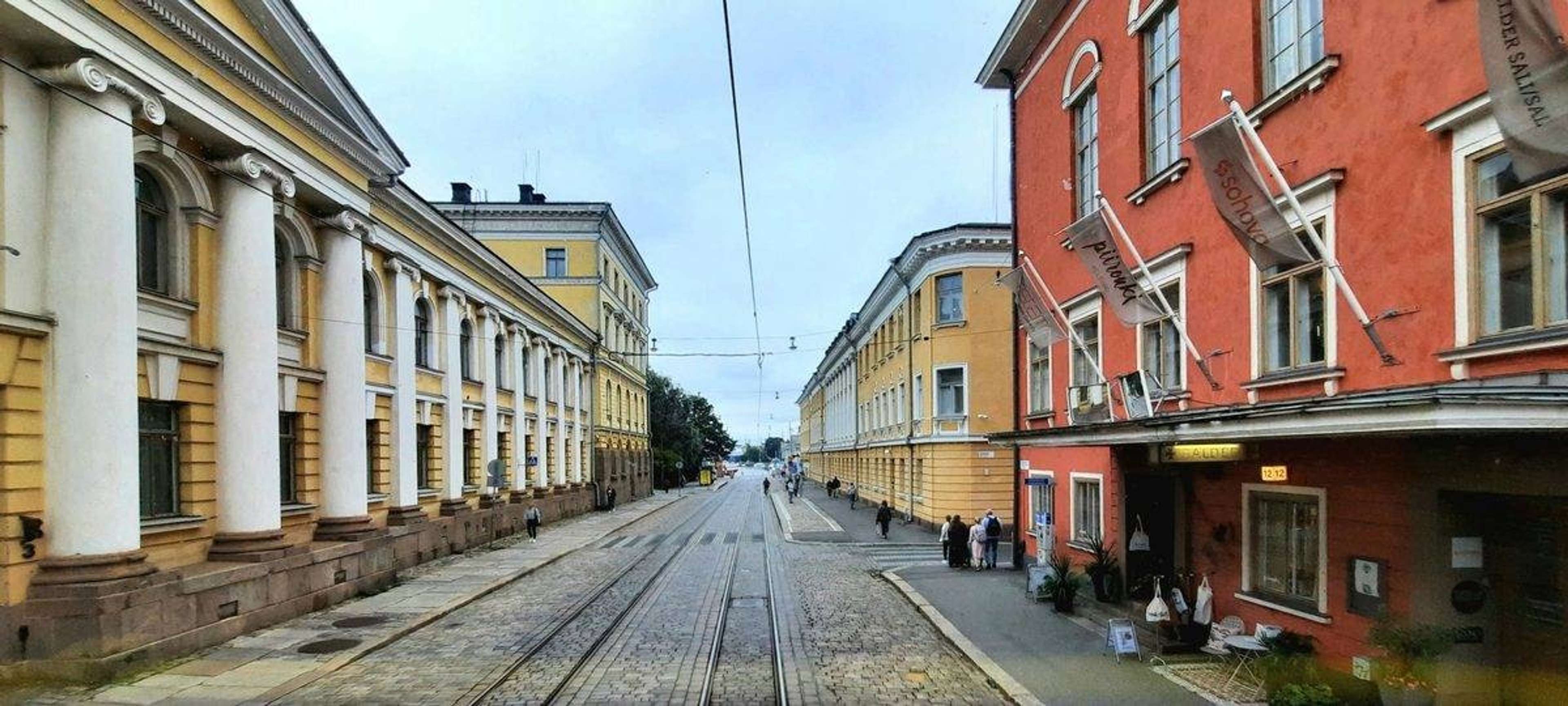 Mukulakivikatu ja raitiovaunukiskot Helsingin keskustassa, sivuilla keltainen pylväsrakennus ja punainen talo