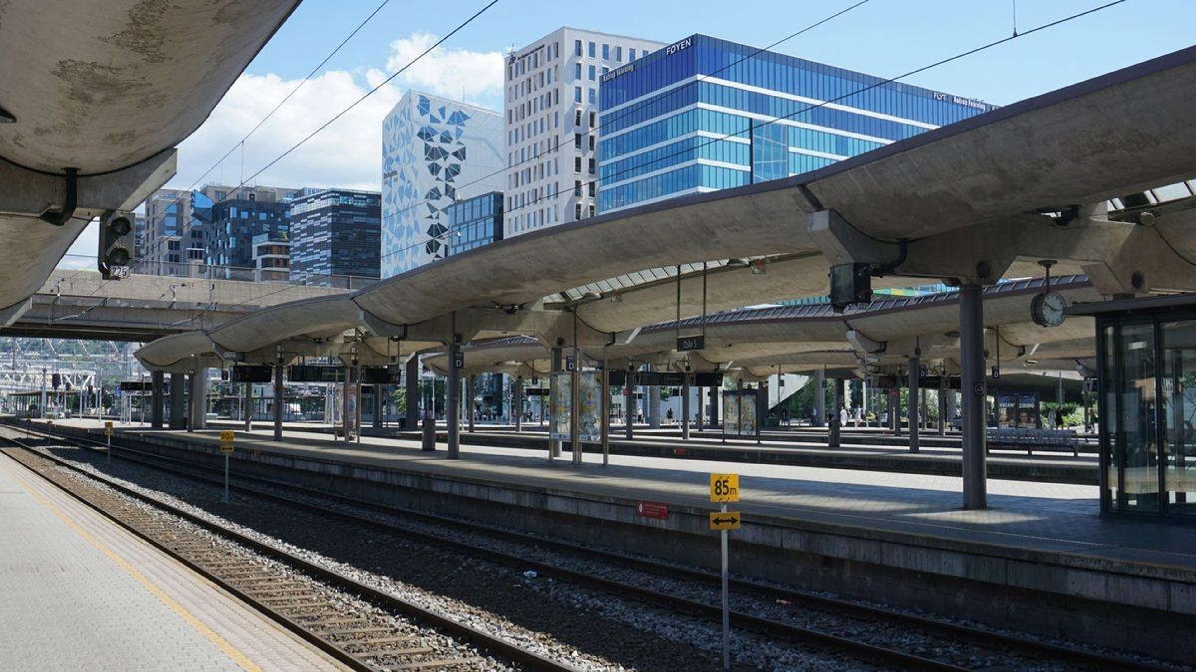 Tomme perronger på en jernbanestasjon med betongtak, skinner og moderne kontorbygg i bakgrunnen
