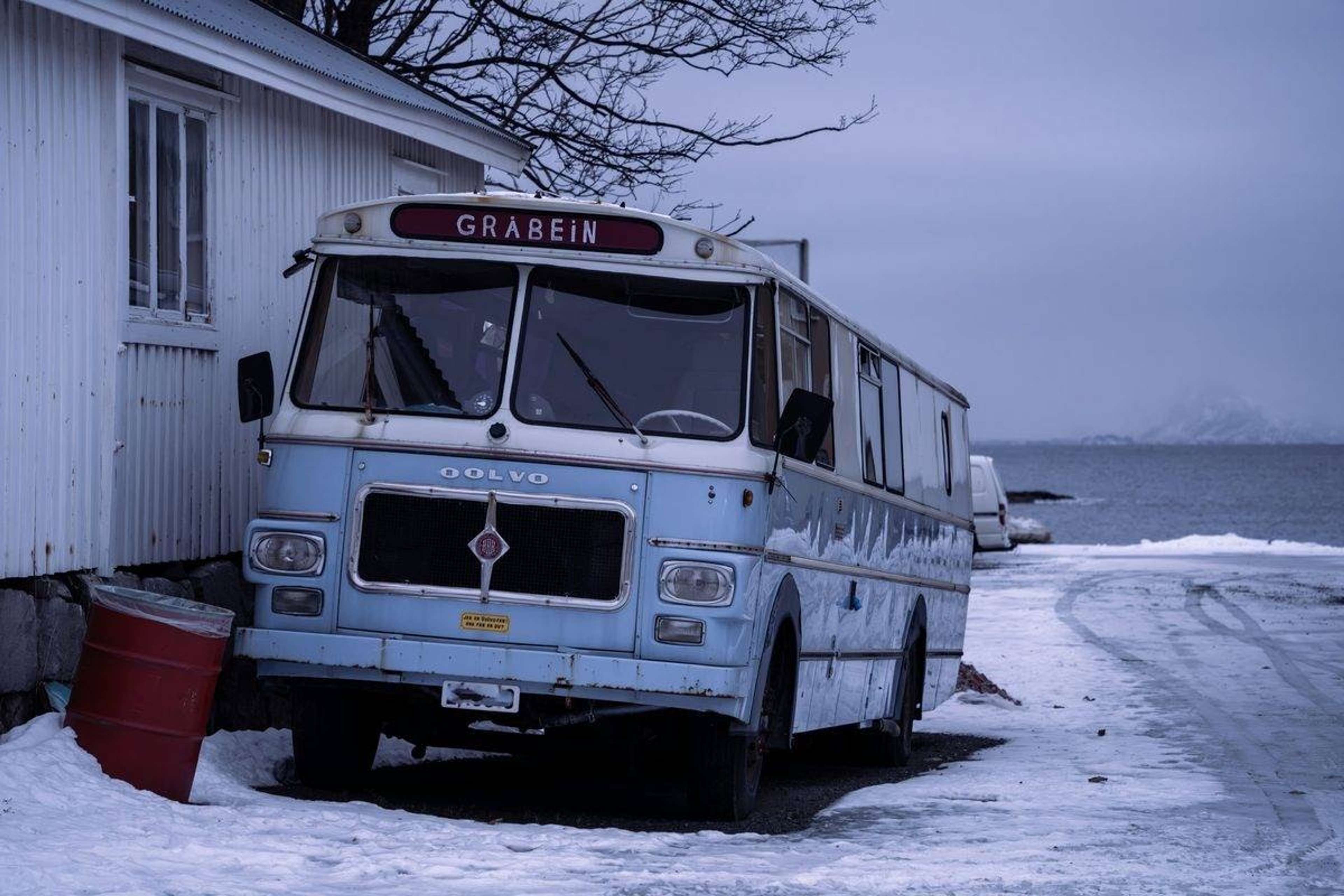 Gammel lyseblå buss parkert i snøen ved et hvitt trehus, med sjø og fjell i bakgrunnen