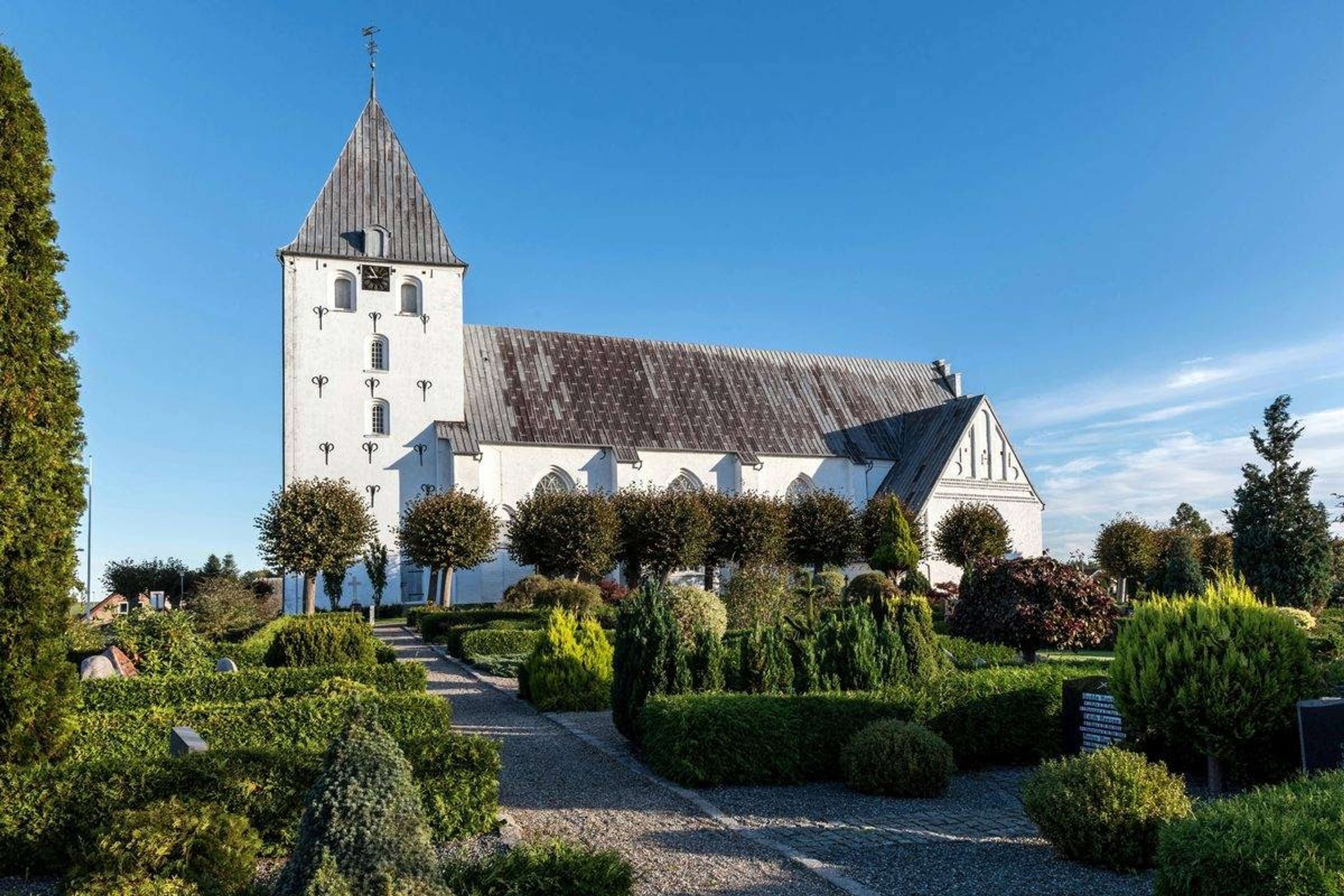 Hvid landsbykirke med gråt tag og klippede hække en solrig dag