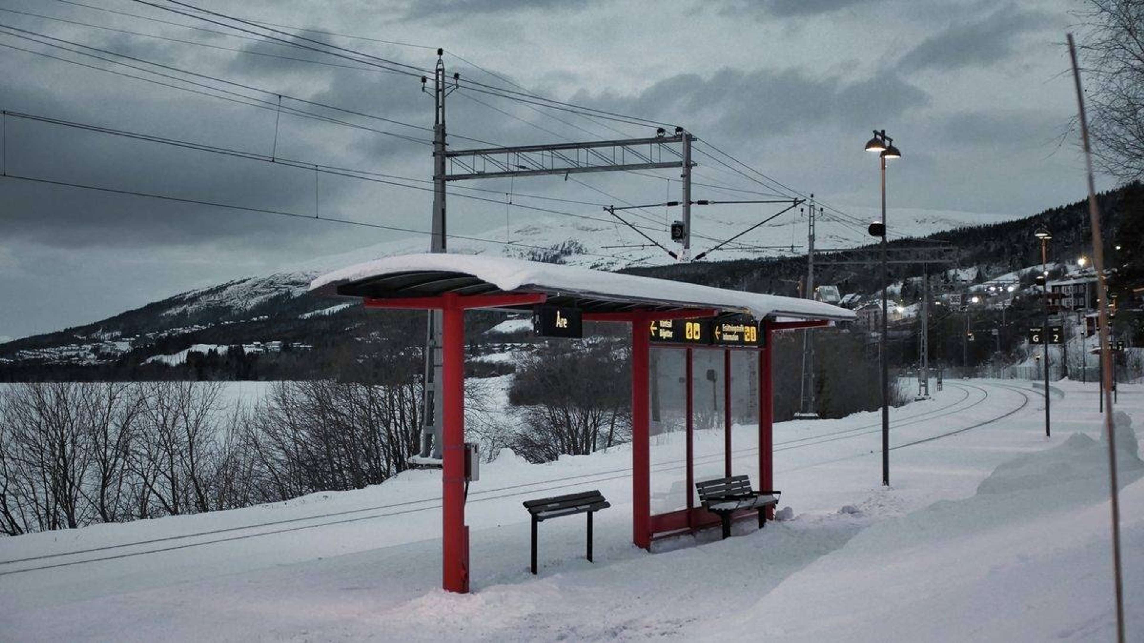 Snöig tågperrong med rött väderskydd och bänkar, spåren svänger bort mot fjällen i skymningen