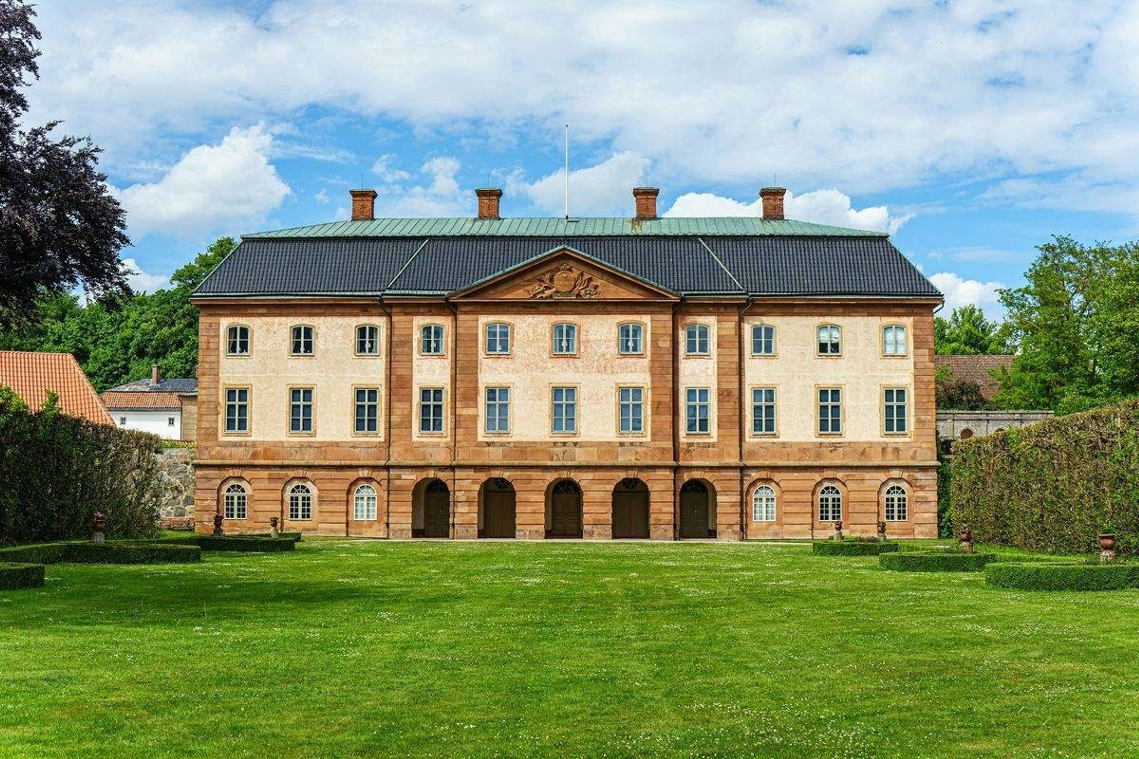 Herrgård i sandfärgad sten med stor gräsmatta framför, blå himmel
