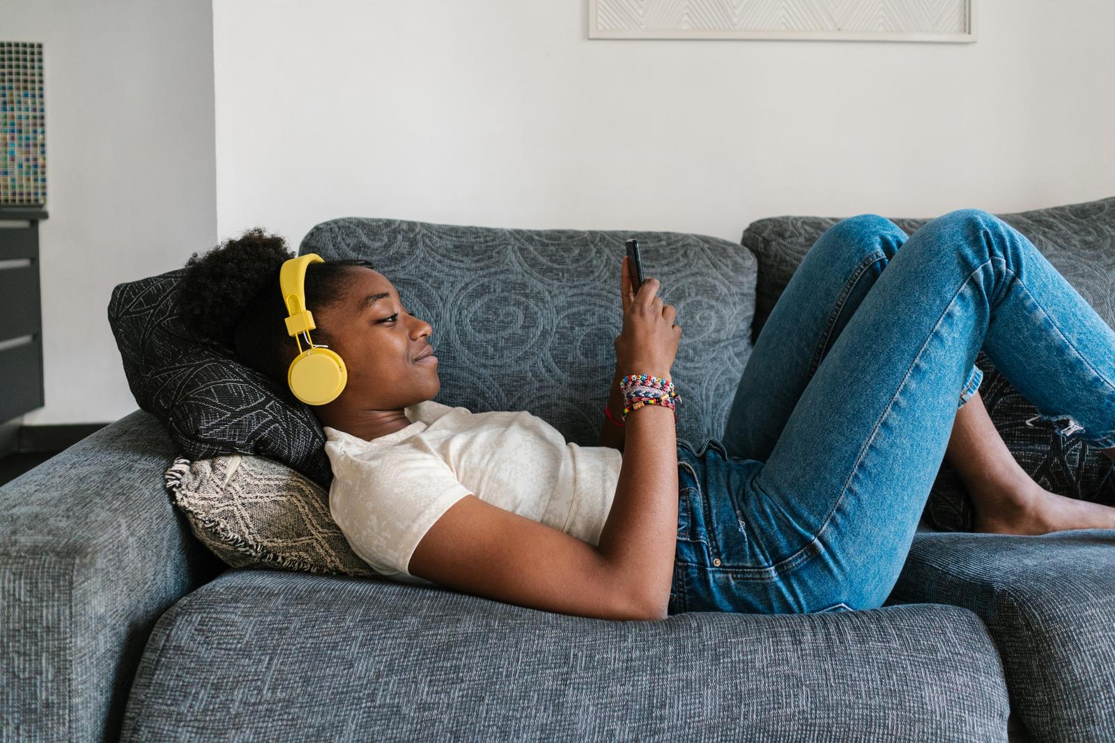 Young woman laying on couch with headphones on scrolling on her smart phone