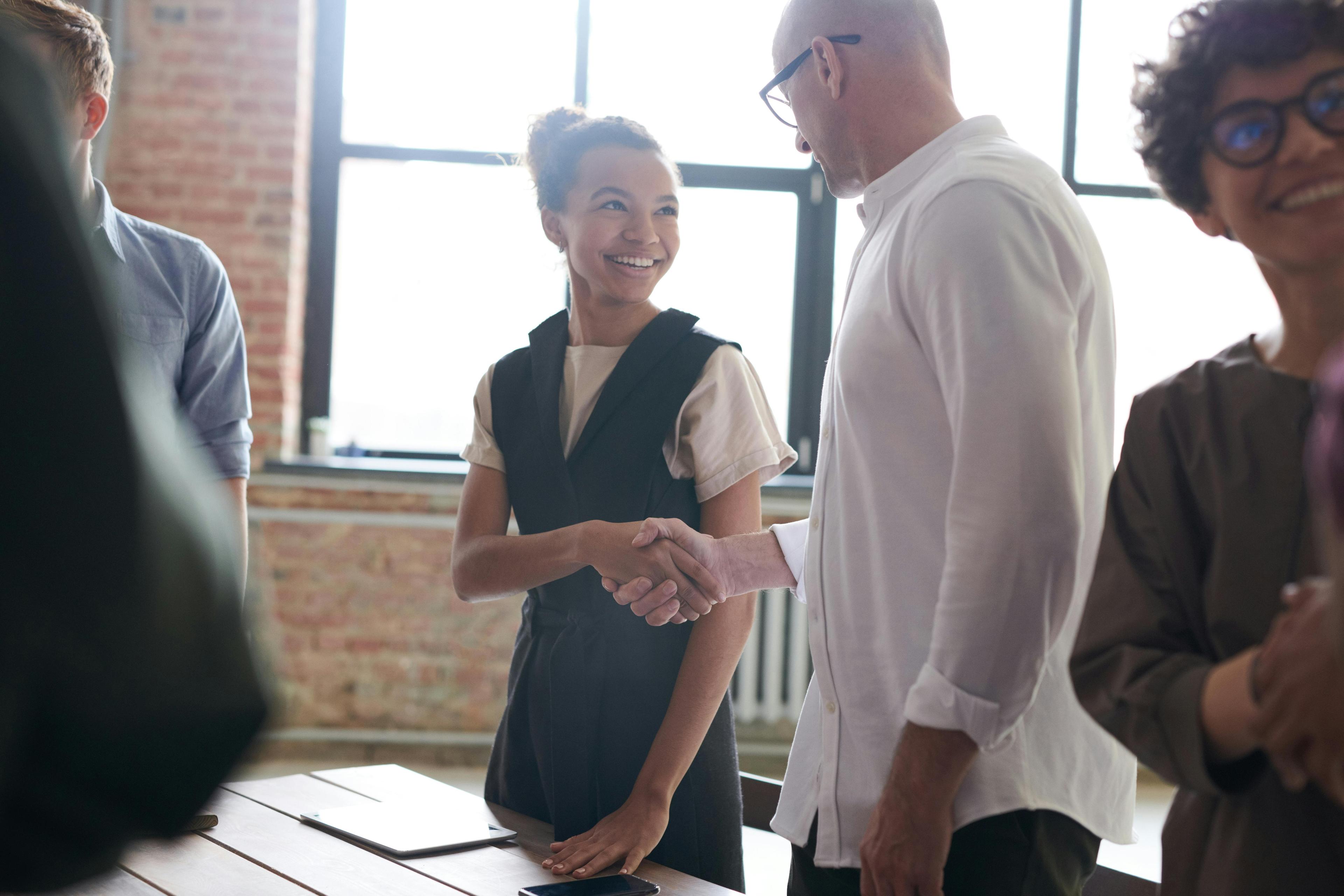 A woman with her hair in a bun shakes hands with a man wearing glasses and a white shirt in a room with other people in front of a window.