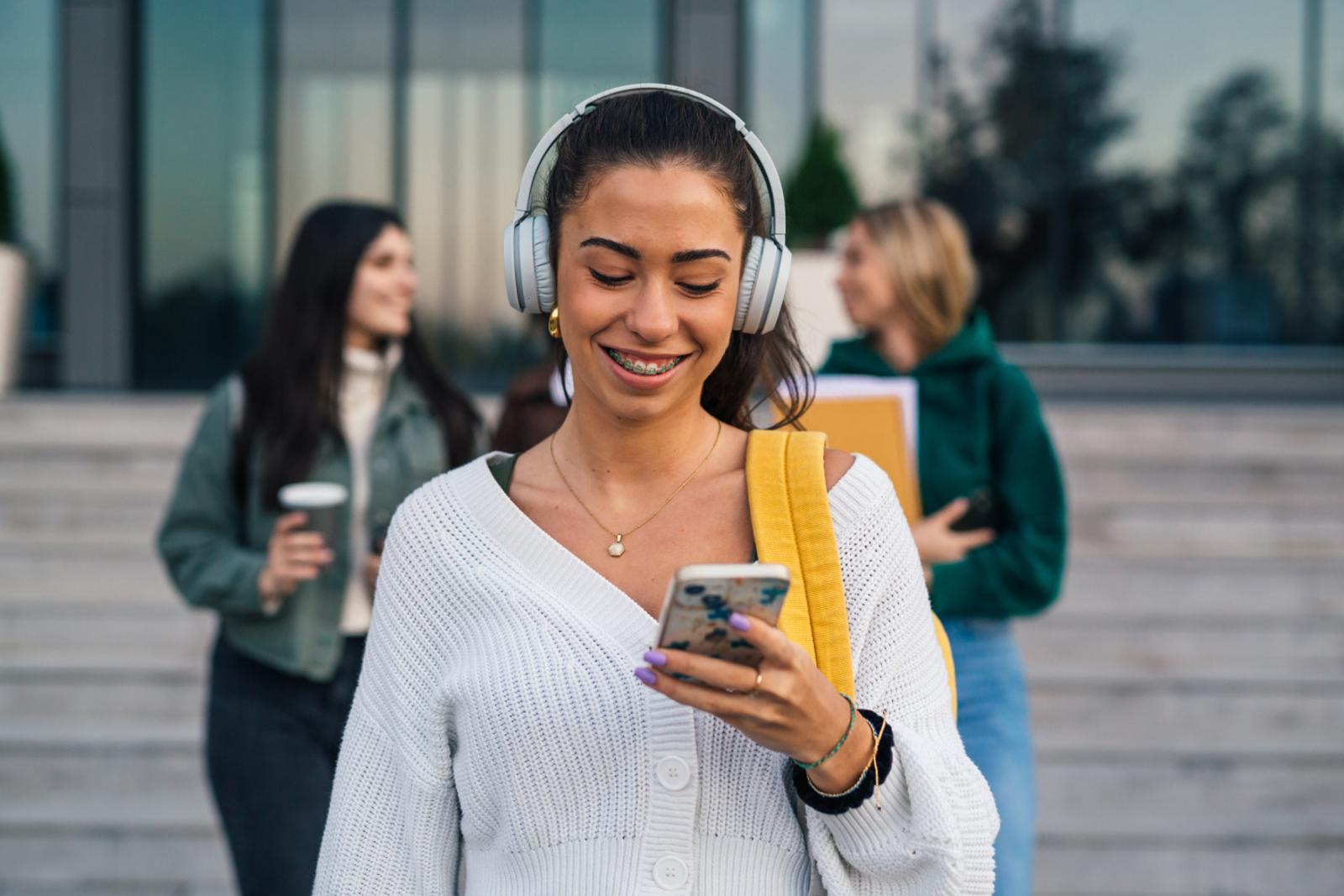 smiling college student wearing headphones while looking at cell phone