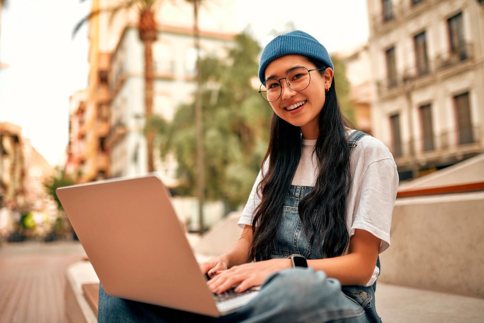 smiling student in overalls and glasses uses laptop on steps of a city library