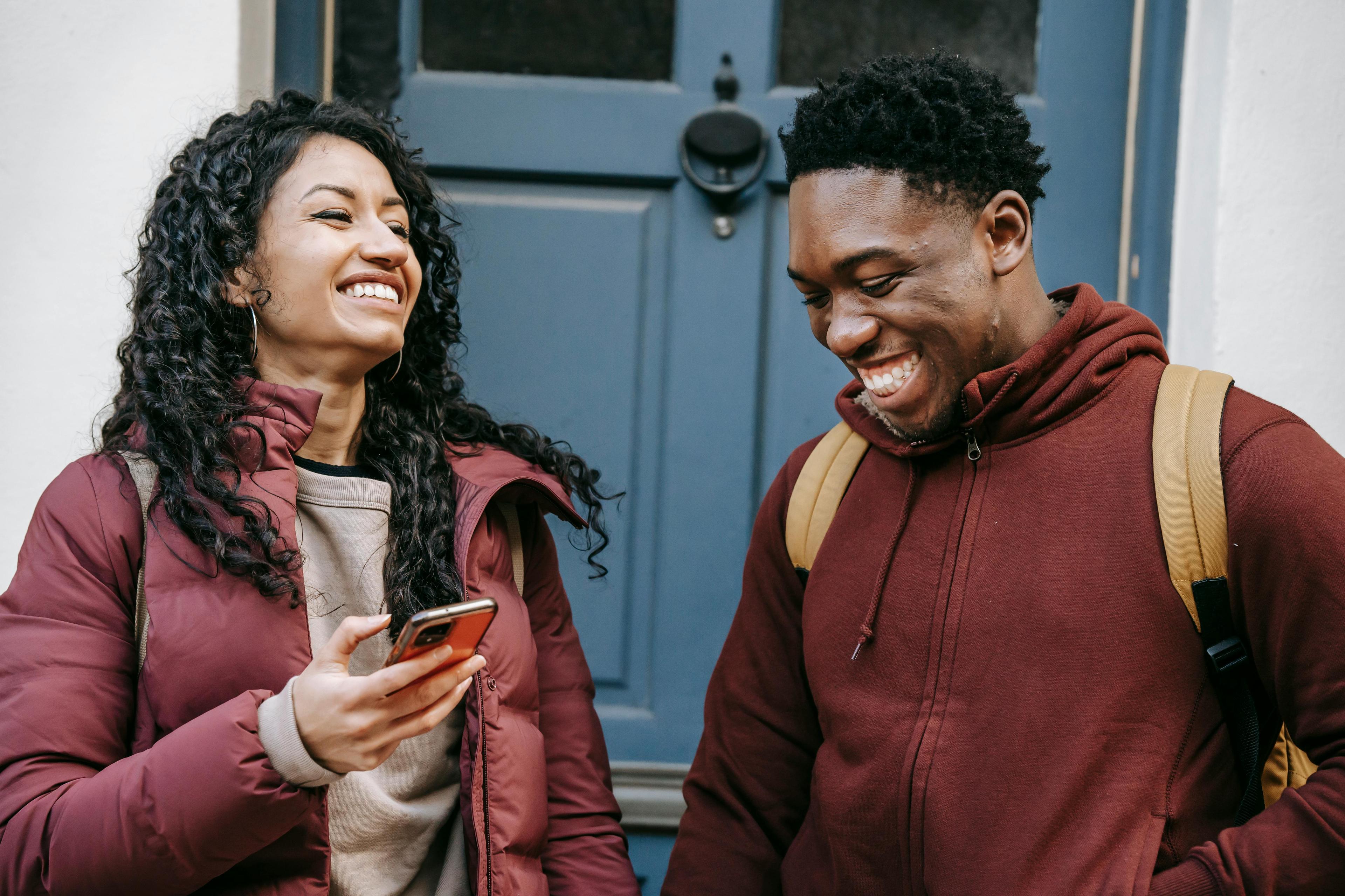 cheerful friends holding smartphones smile while standing near door to a building