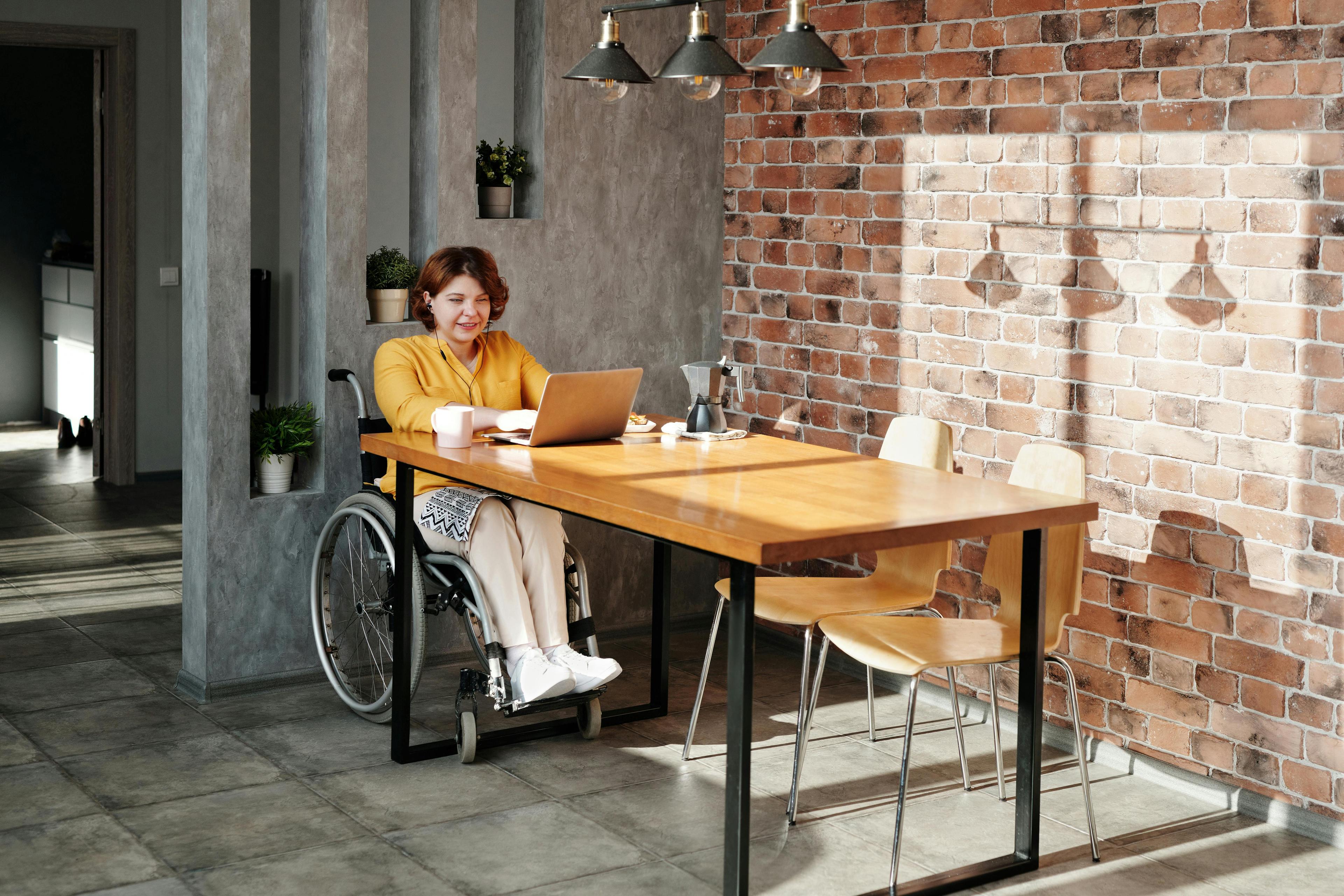 A young woman sitting in a wheelchair in a cafe using her laptop. She has a small smile and appears optimistic about what she sees on the screen.