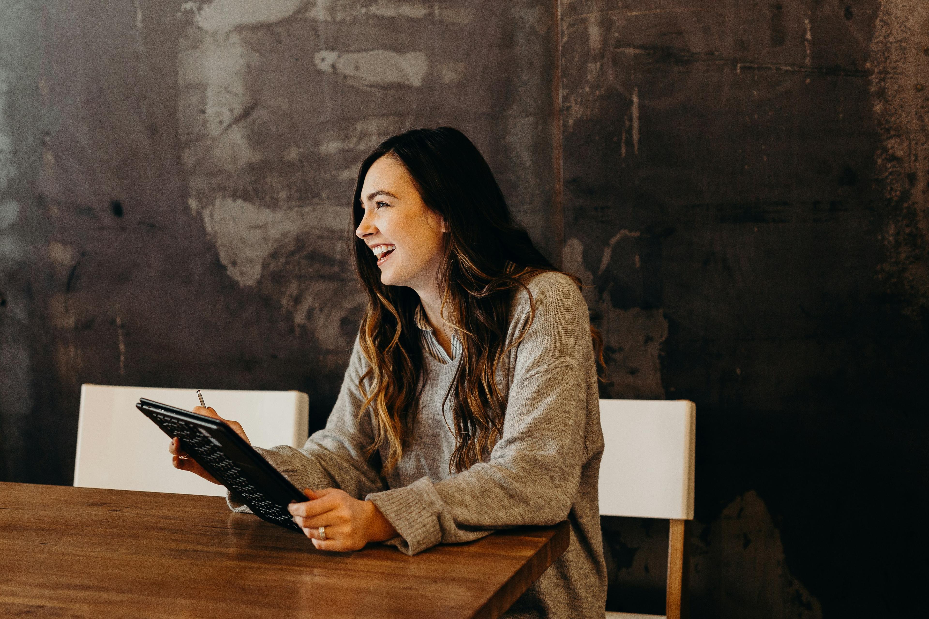 A white woman with long brown hair in a heather gray sweater sits at a desk holding an iPad in front of a dark gray wall.