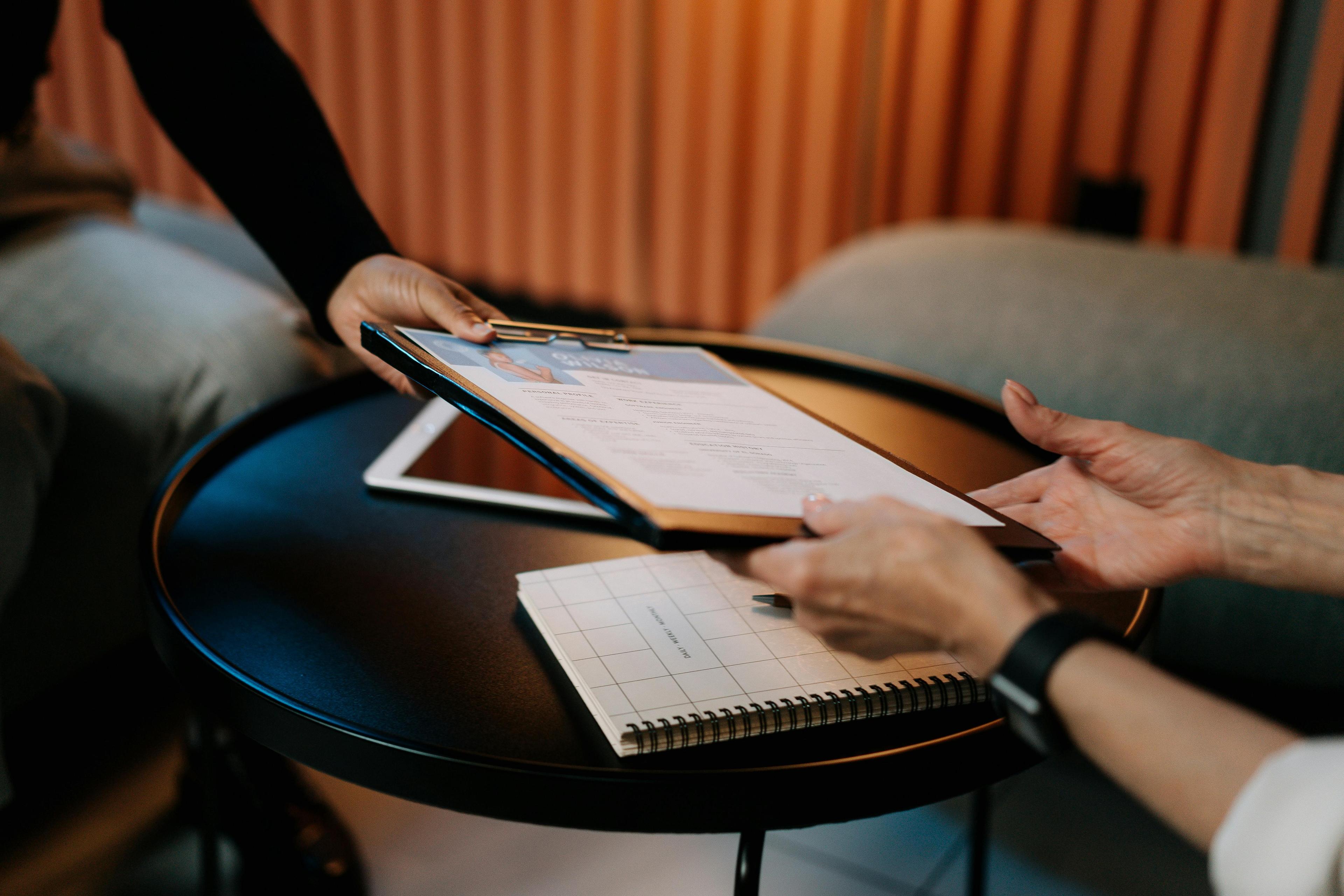 An arm in a black long sleeve top hands a clipboard to two awaiting older hands, in front of a table and orange back drop. 