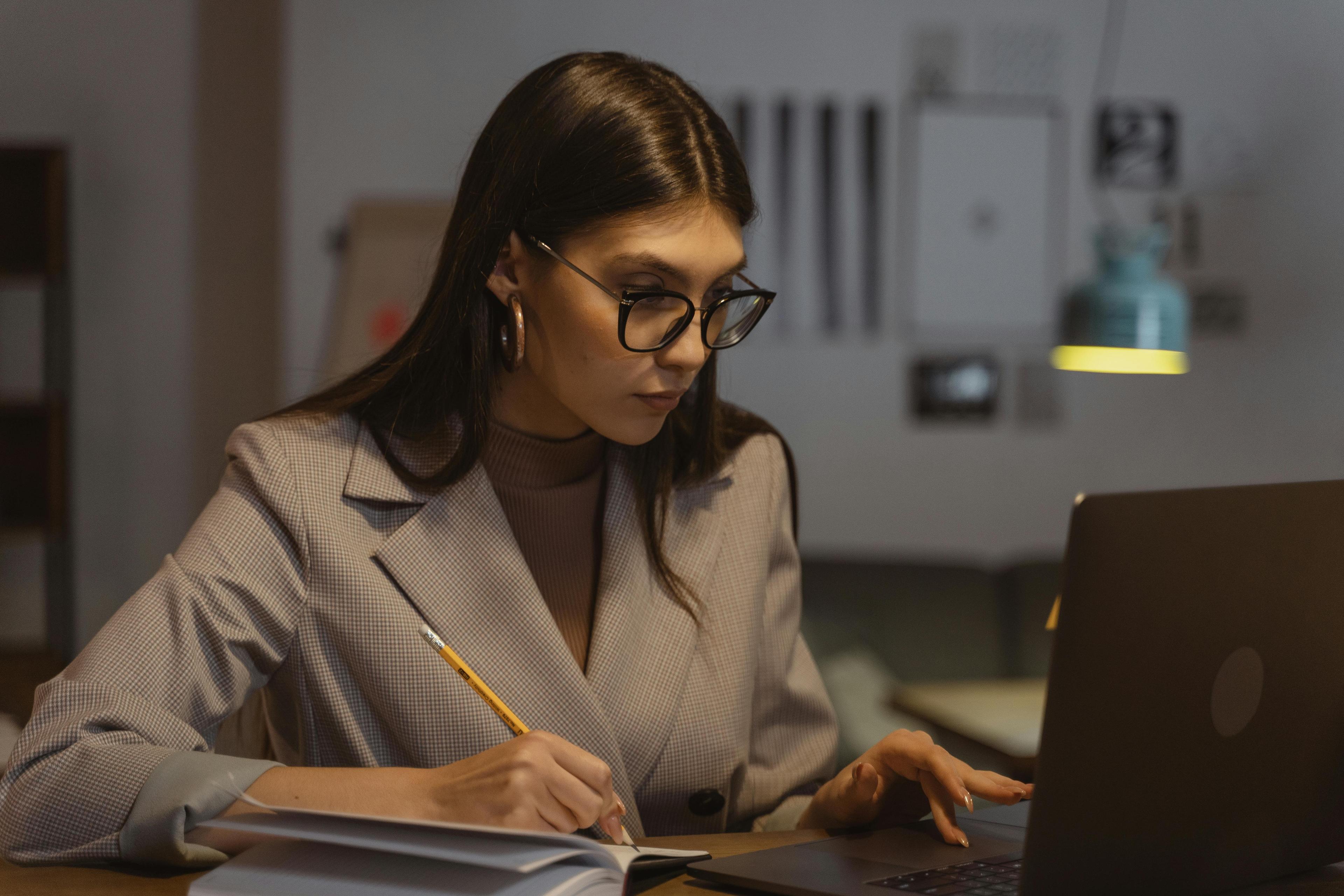 A woman with dark brown hair and glasses wears a gray blazer while she sits in front of a computer taking notes with a pencil in a notebook.