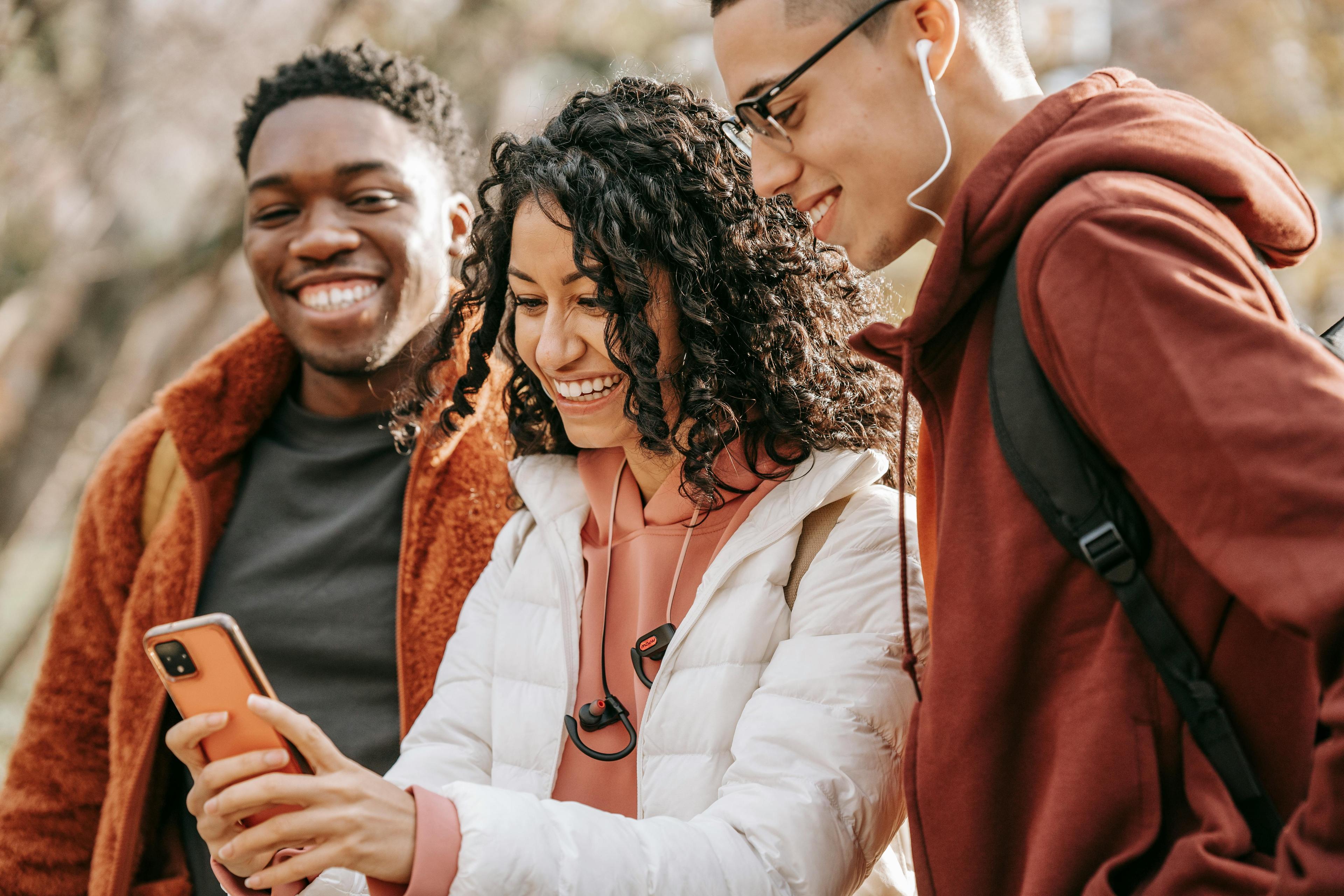 Two men in orange sweatshirts stand next to a woman with curly hair in a white puffer jacket outside looking at a cellphone together.