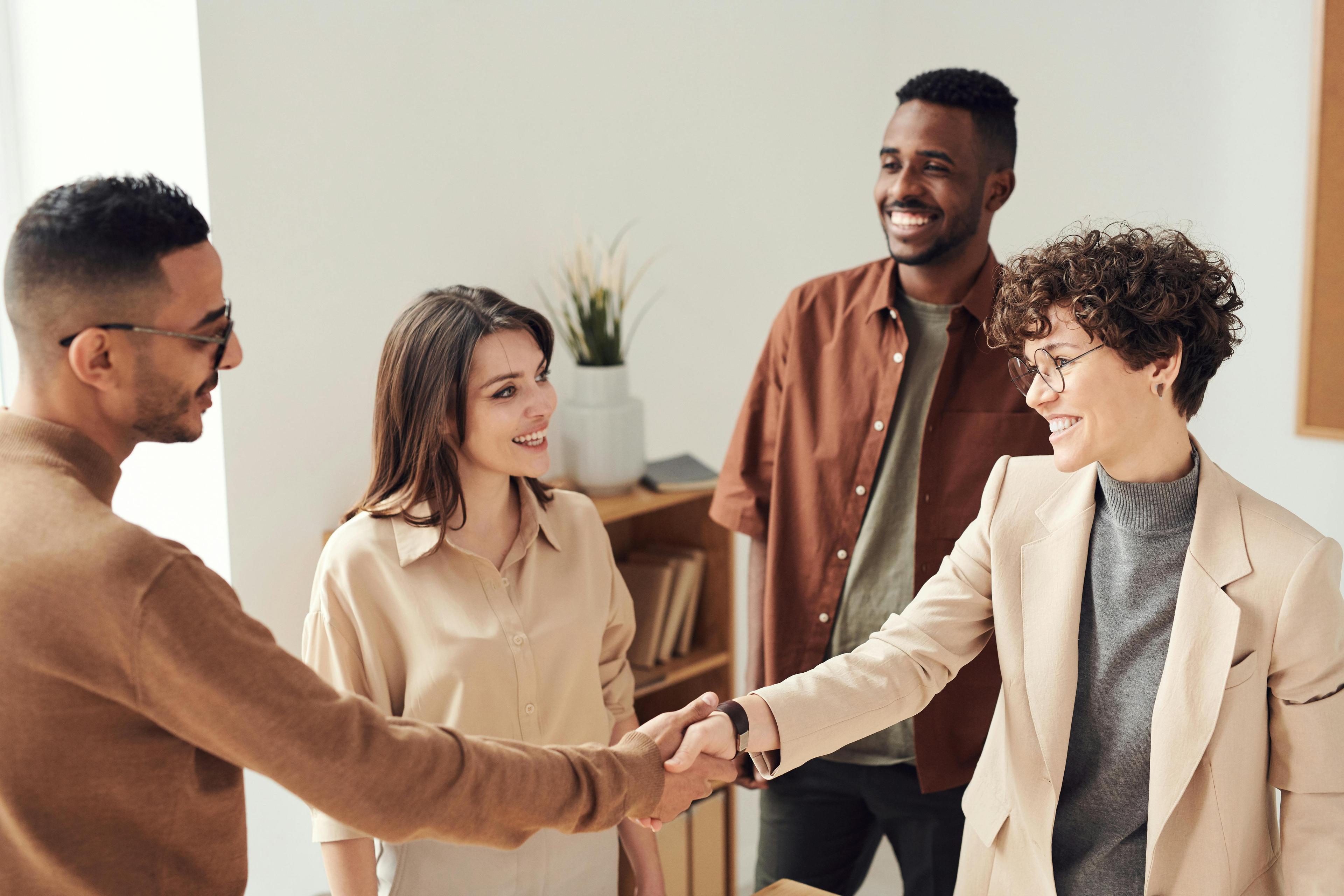 A man in a light brown turtleneck and a woman in a cream blazer shake hands in front of a different man and woman. They all stand in a bright room with white walls.