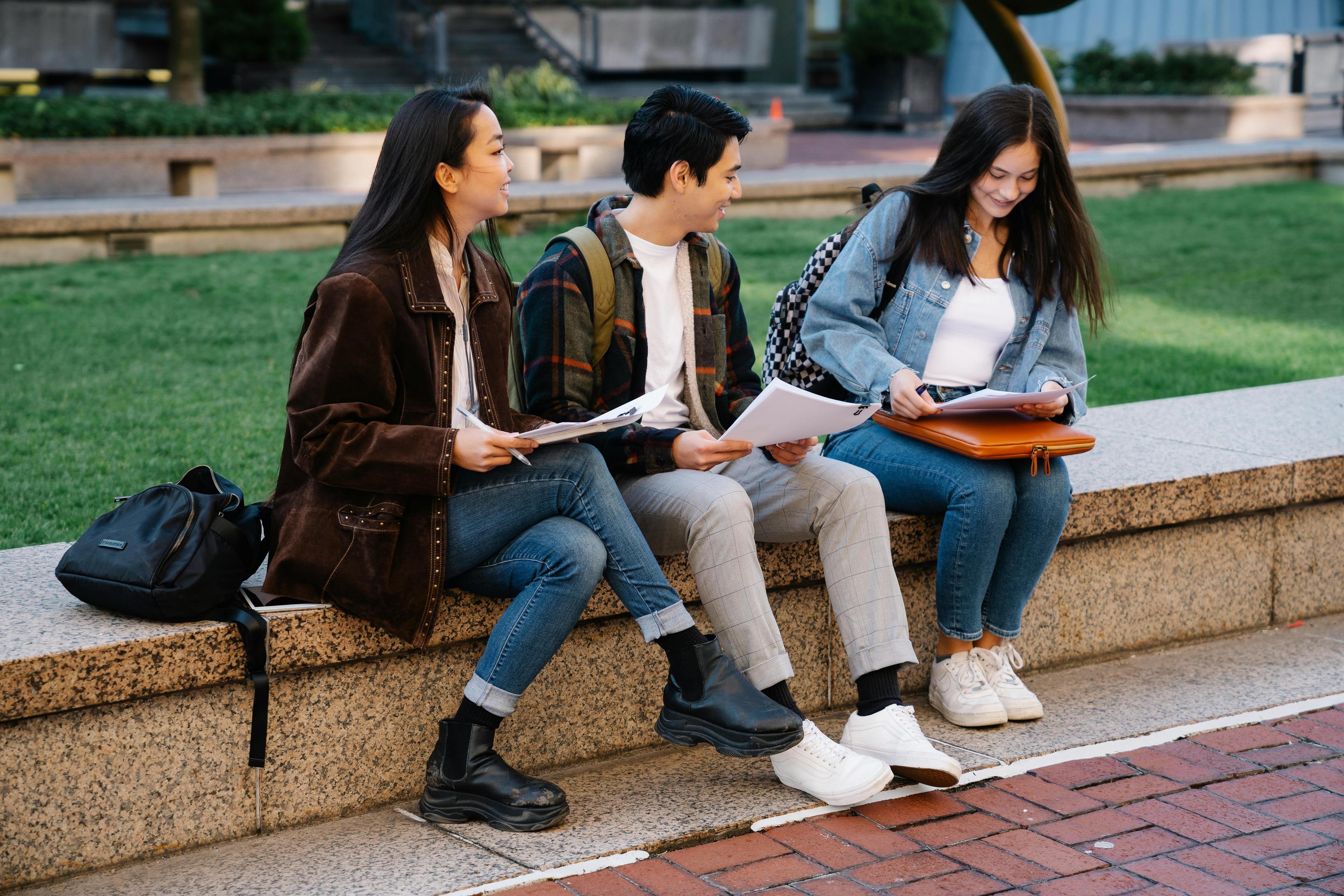 A young man sits between two women on a short ledge outside in front of grass. They all have backpacks and papers.