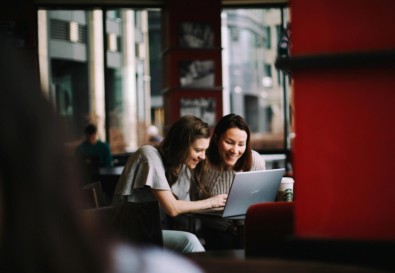 Two women sitting at a café, smiling and looking at a laptop together, with coffee cups on the table and a cityscape visible through large windows in the background.