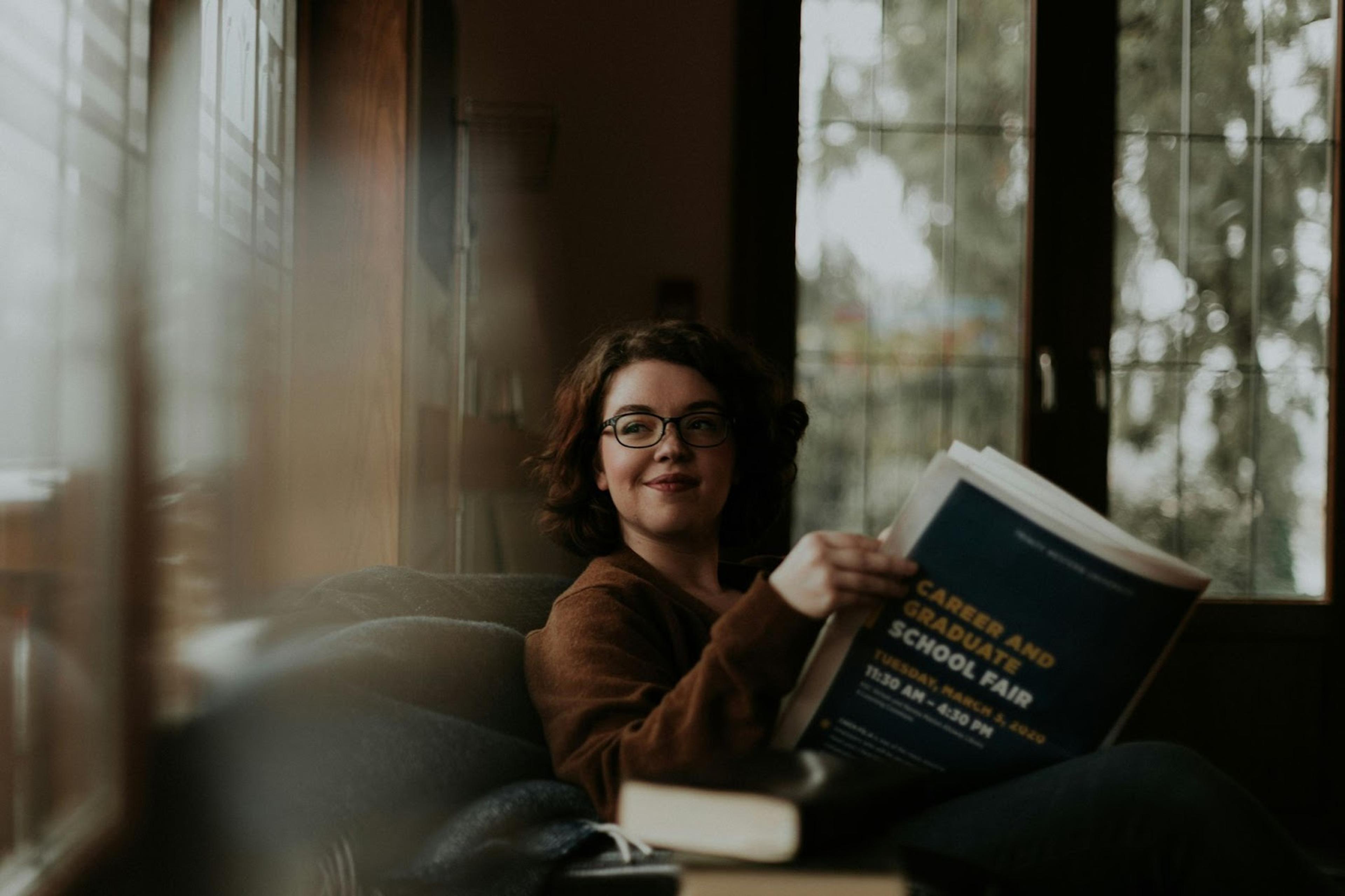 A white woman with short brown hair and glasses in brown coat sits in front of windows and reads a "career and graduate school fair" brochure