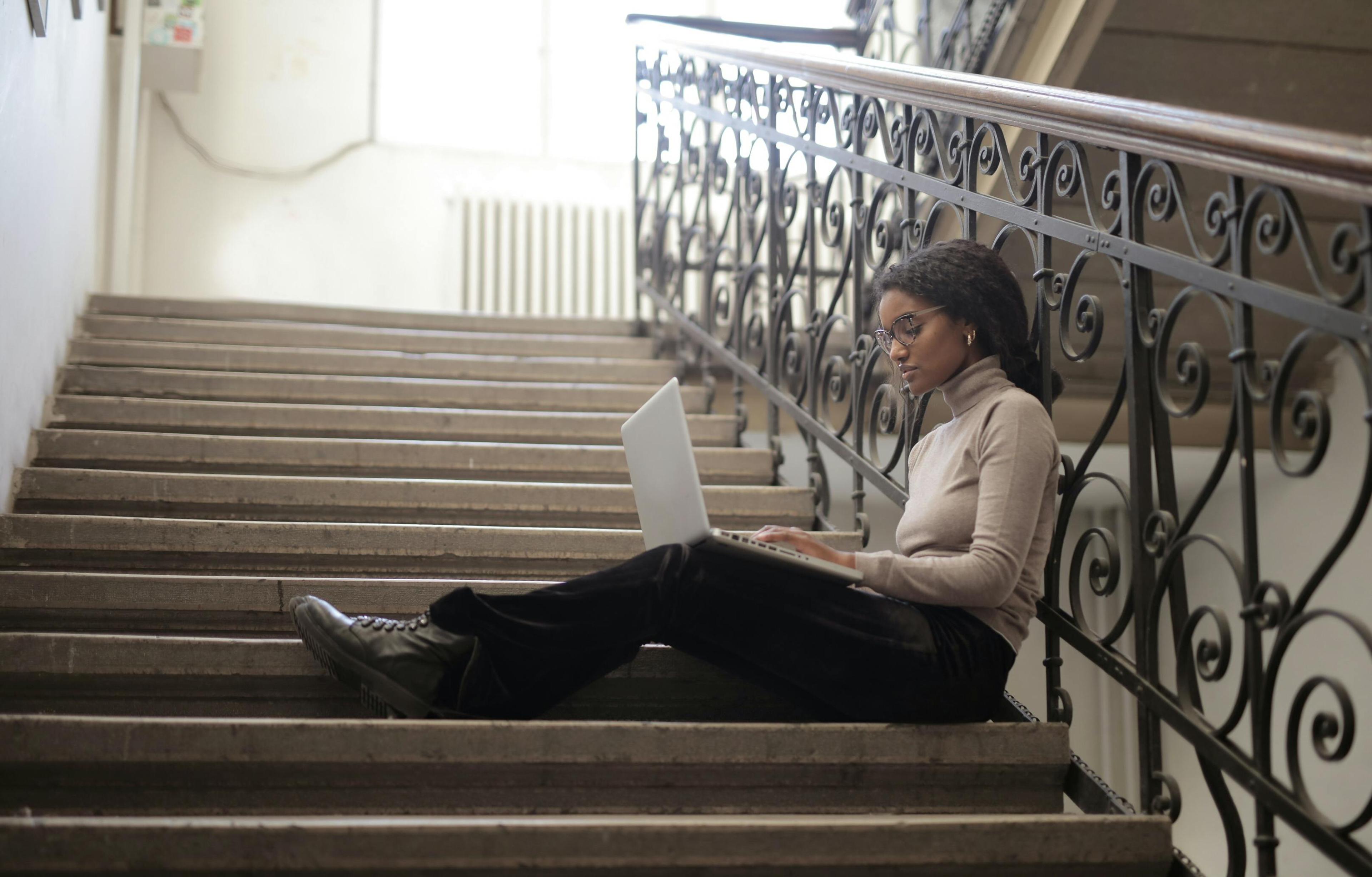 a black woman types on her computer while sitting on a staircase in a stone colored turtleneck and black pants