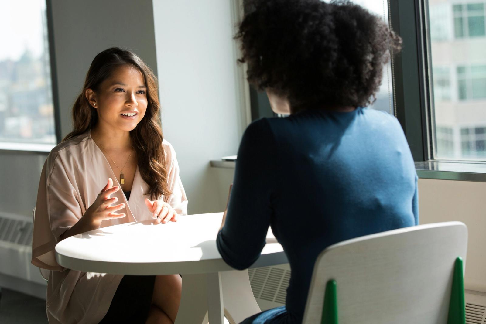 Two women sit across from each other at a small round table in an office, engaged in conversation.