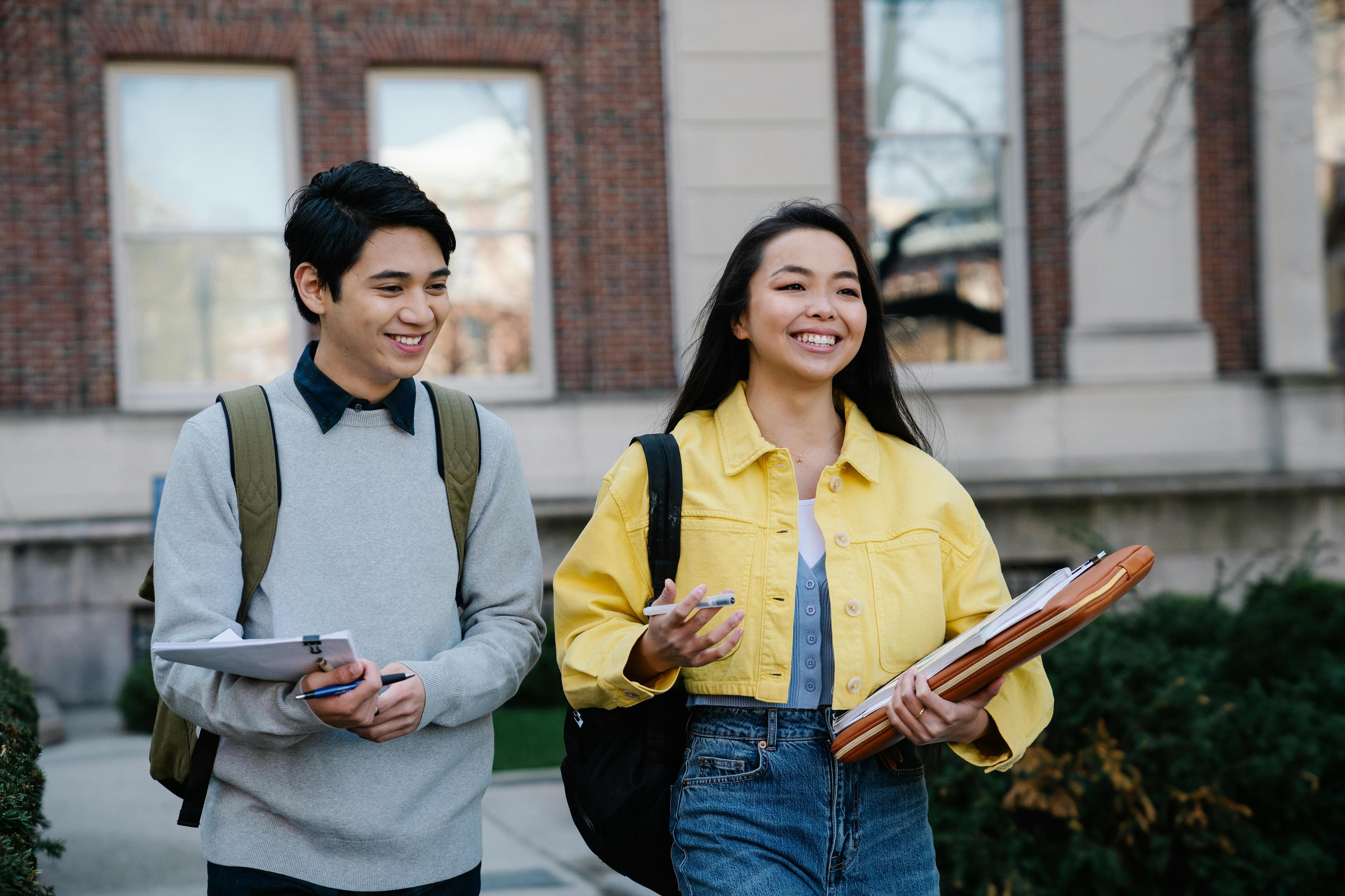 A man with short brown hair and a grey sweater walks outside a building alongside a woman with long black hair, who is wearing a bright yellow cropped jacket. Both are holding papers, and she is holding a laptop case.