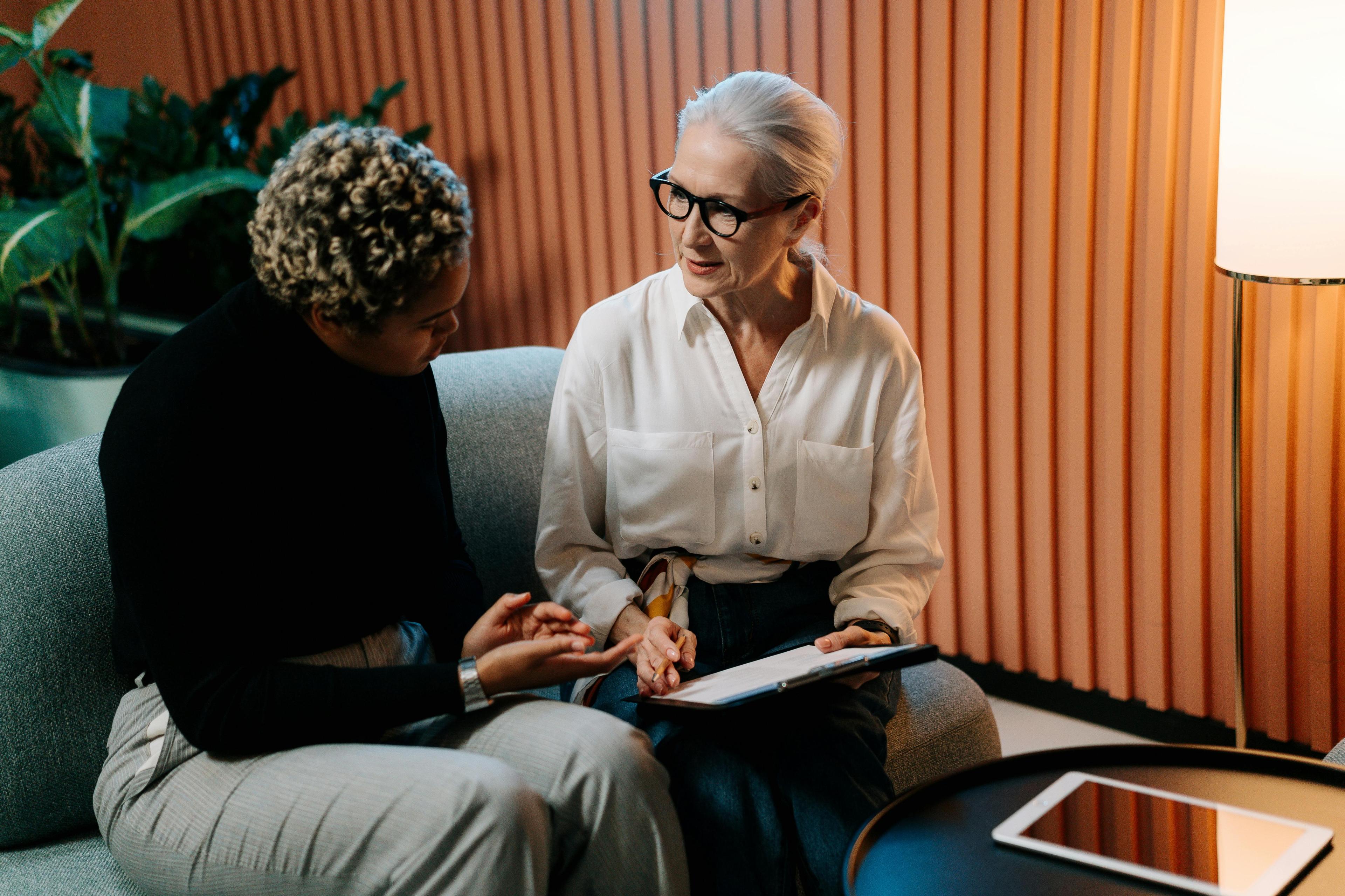 a white woman holding a clipboard sits next to a black woman on a couch in front of a textured orange wall.