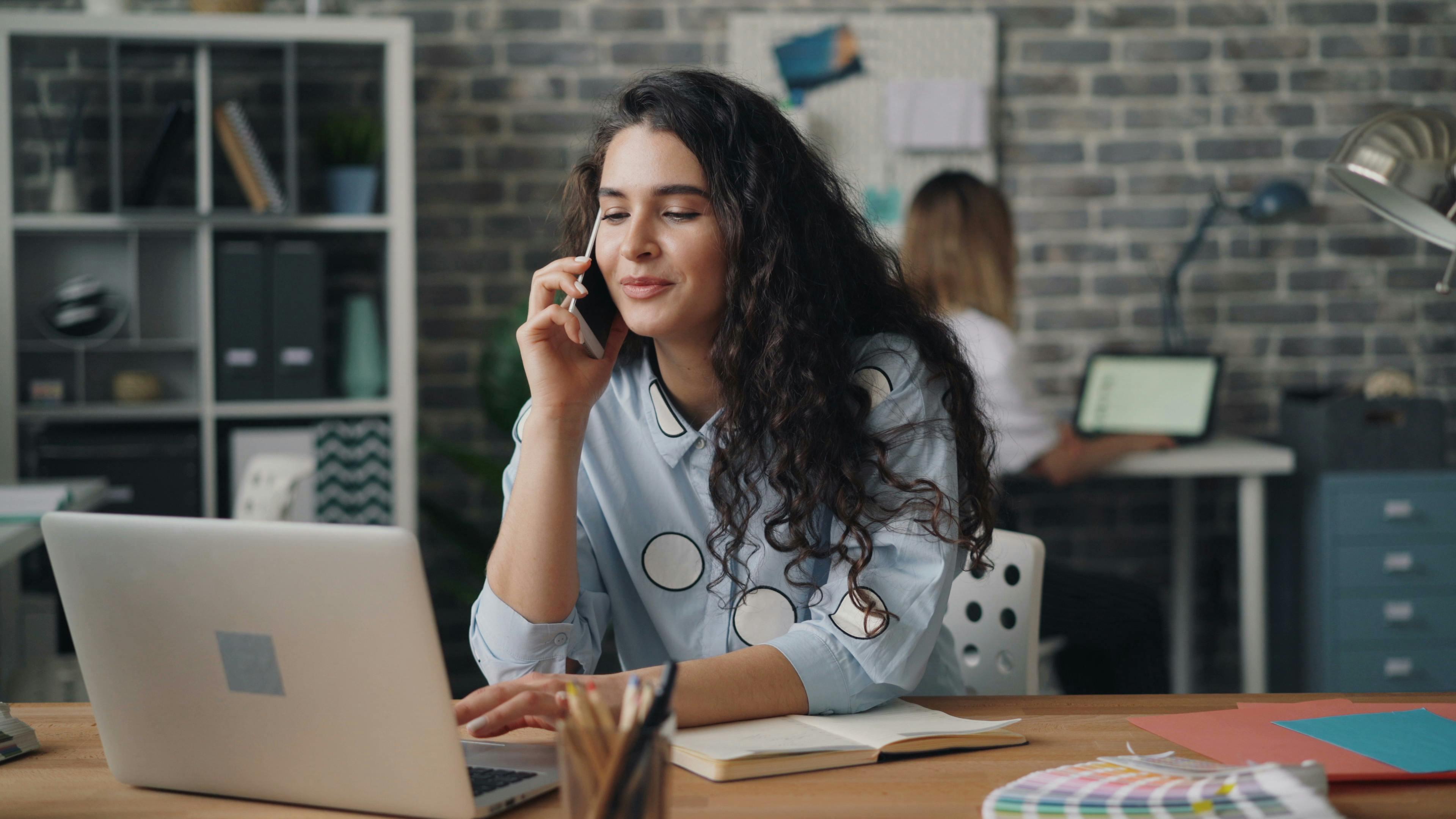 woman with long curly brown hair holding cellphone to her ear while typing on a laptop in front of a workspace with a brick wall