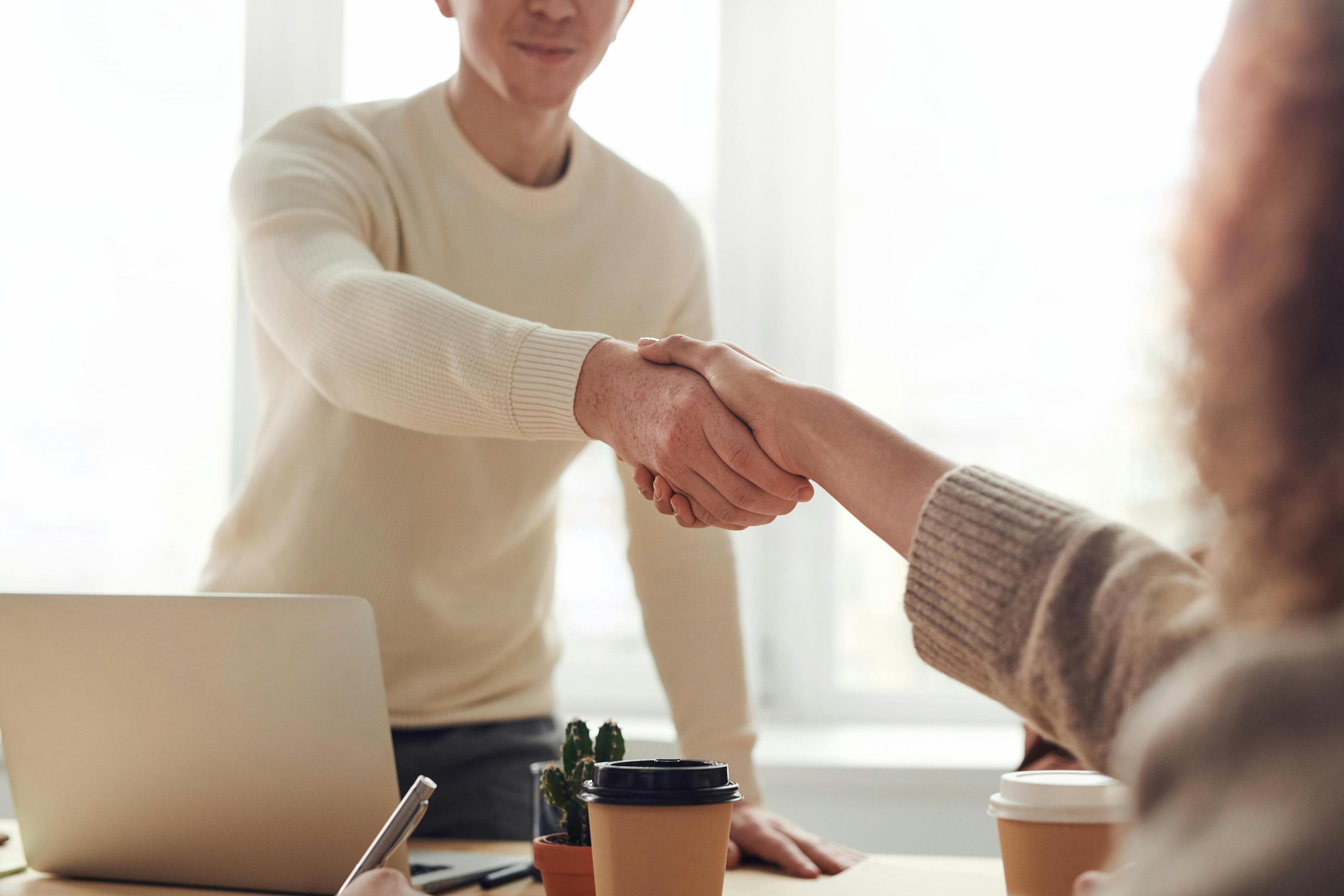 A person in a cream sweater shakes the hand of another person in a brown sweater over a desk in front of a bright window.