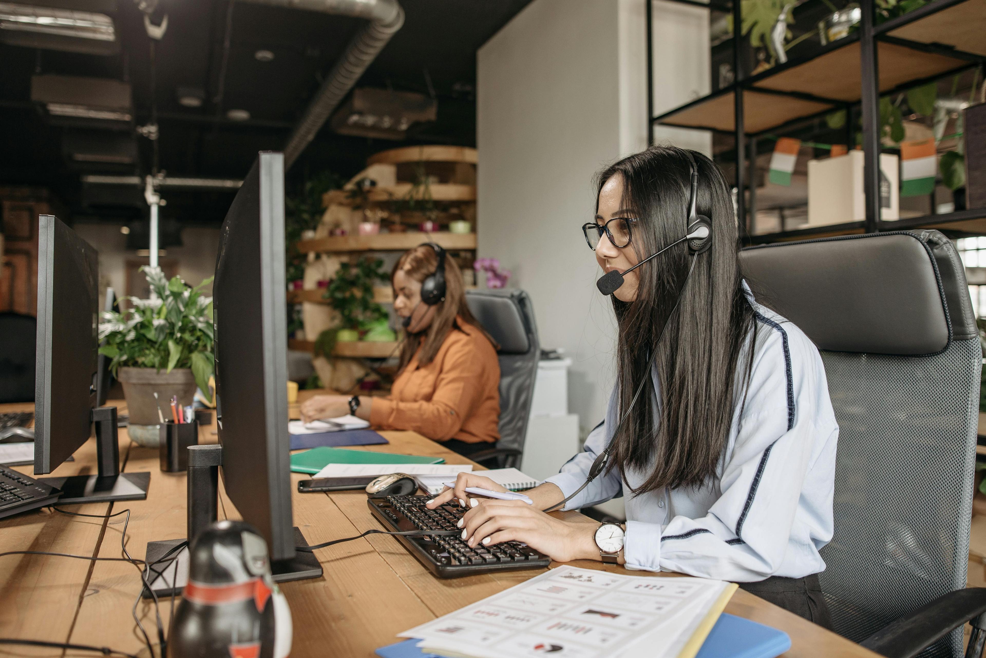 Two women sit behind a long desk looking at desktop computers with headsets on.