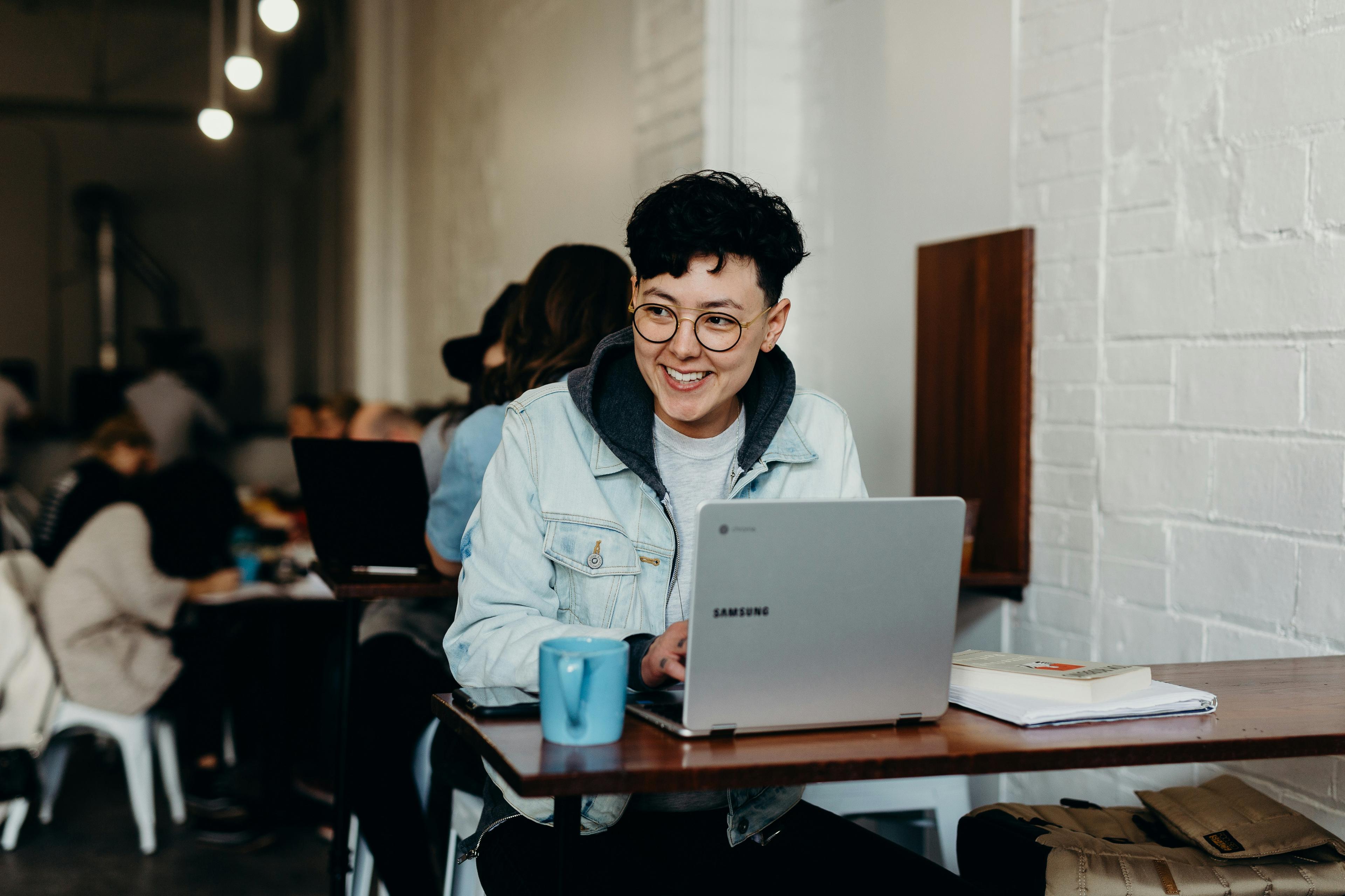 A person with short curly dark brown hair and glasses sits at a table behind a laptop and smiles. They are against a white brick wall.