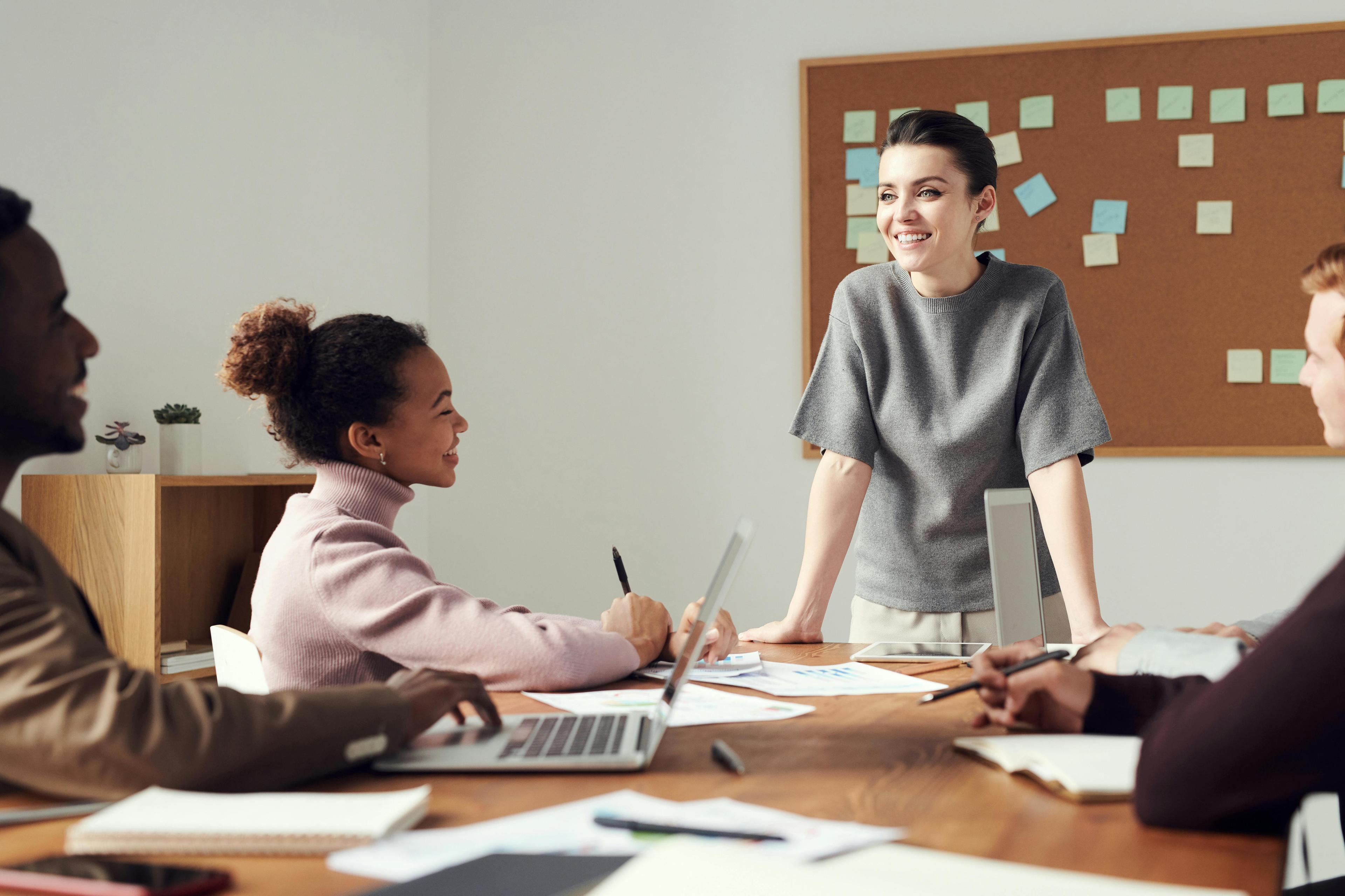 Two men and a woman sit at a table with papers and a laptop, they are all looking at a woman standing at the head of the table in front of a cork board covered in sticky notes.