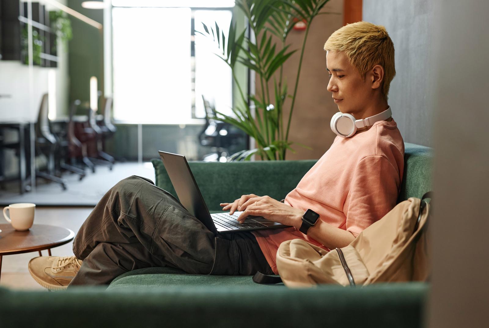 Young professional working remotely on a laptop in a modern coworking space, wearing casual clothes and headphones, sitting on a green couch with natural light and plants in the background.