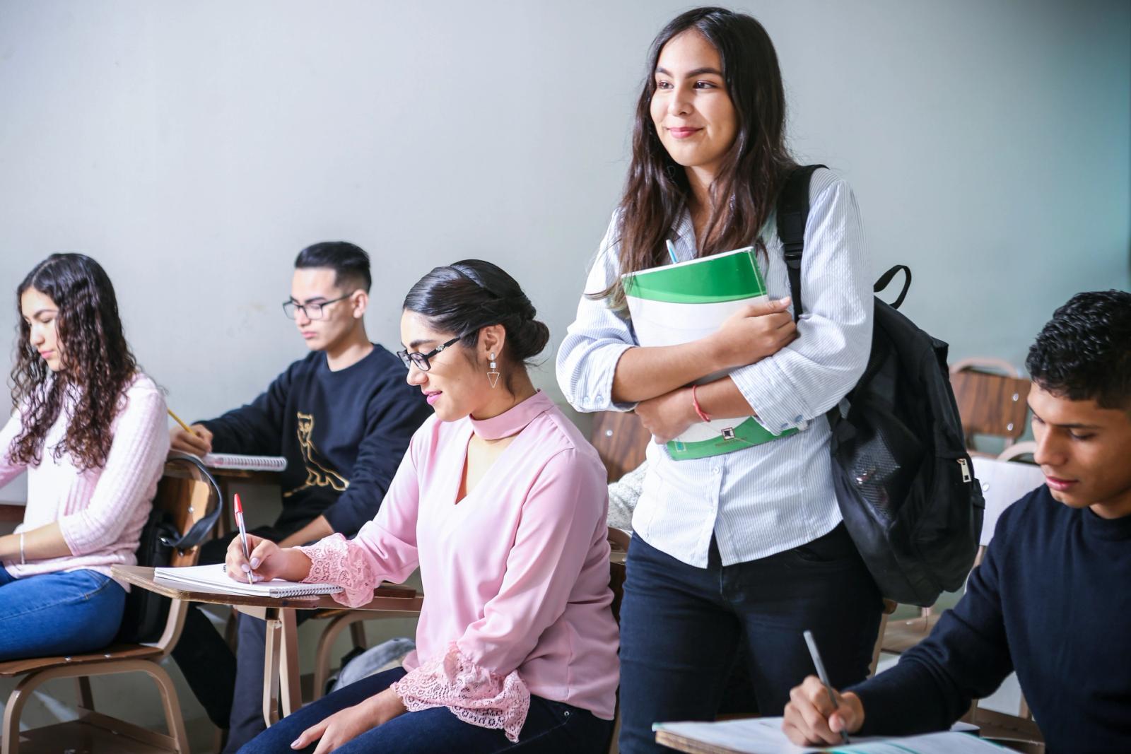 Girl standing in classroom holding books while everyone else studies