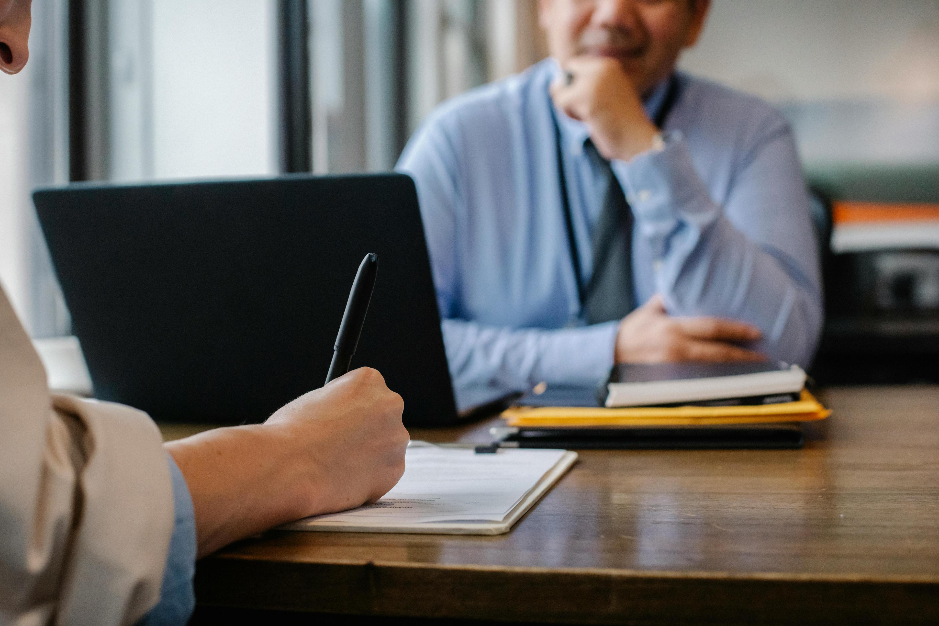 Person's arm signing document on a brown desk across from a man in a blue shirt