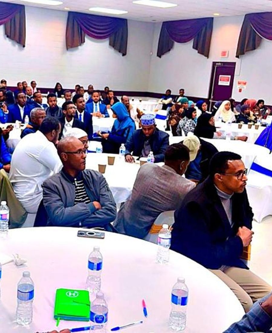 A packed hall of community members seated at round tables, listening attentively.