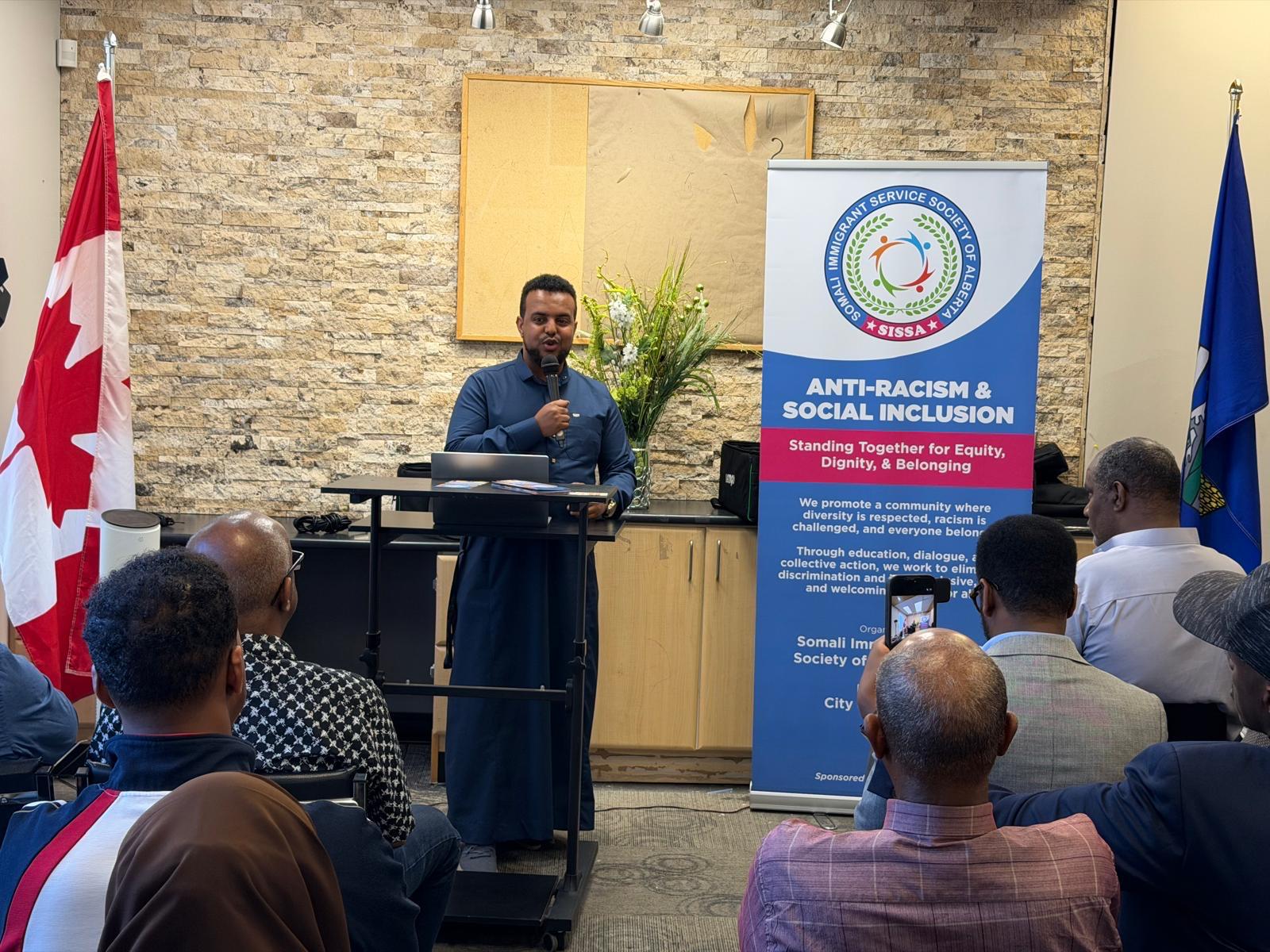 A speaker at a podium addresses attendees, with the SISSA Anti-Racism & Social Inclusion banner and Canadian and Alberta flags behind.