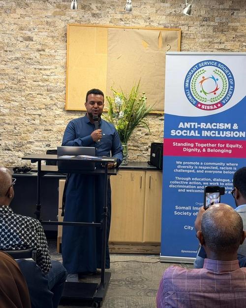 A speaker at a podium addresses attendees, with the SISSA Anti-Racism & Social Inclusion banner and Canadian and Alberta flags behind.