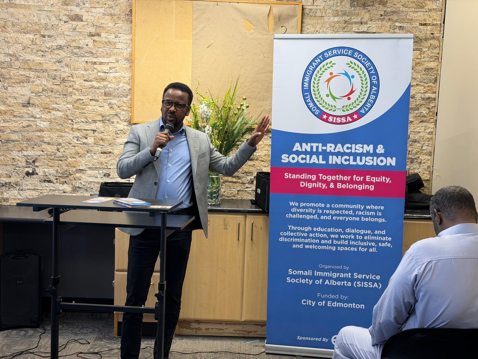 A speaker with a microphone addresses the room beside the SISSA Anti-Racism & Social Inclusion banner, which credits funding from the City of Edmonton.