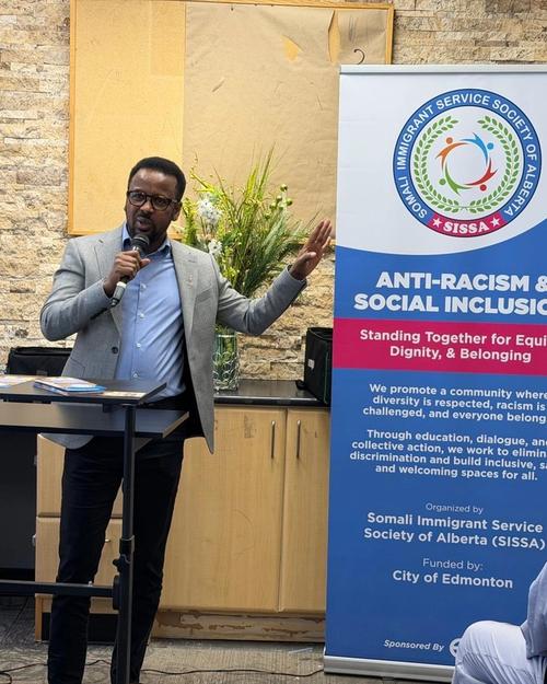 A speaker with a microphone addresses the room beside the SISSA Anti-Racism & Social Inclusion banner, which credits funding from the City of Edmonton.