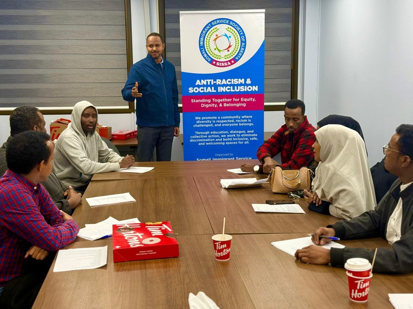 Adults seated around a table with handouts listen as a facilitator speaks beside a SISSA Anti-Racism & Social Inclusion banner.
