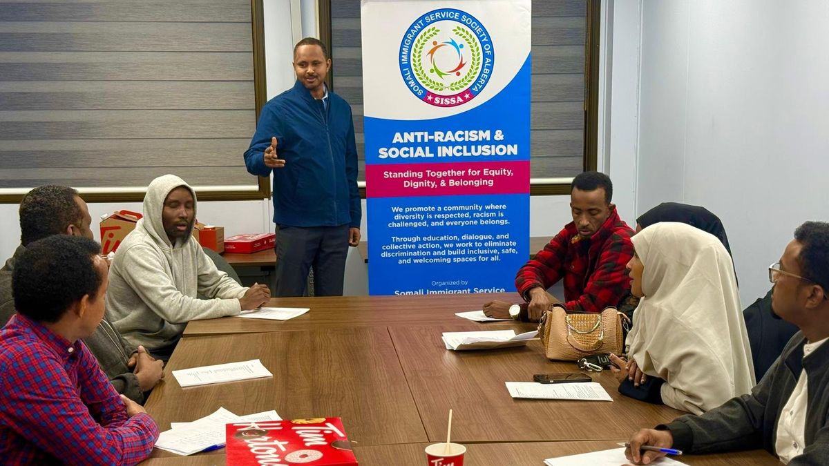 Adults seated around a table with handouts listen as a facilitator speaks beside a SISSA Anti-Racism & Social Inclusion banner.