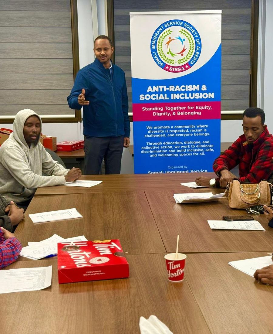 Adults seated around a table with handouts listen as a facilitator speaks beside a SISSA Anti-Racism & Social Inclusion banner.